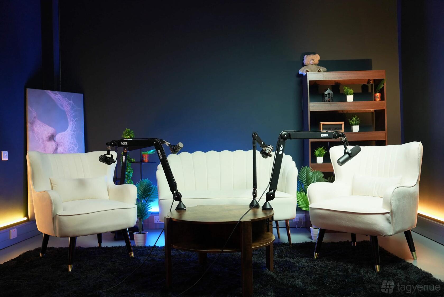 A podcast studio with white armchairs, a curved sofa, microphone arms, and potted plants at NextMedia.London.