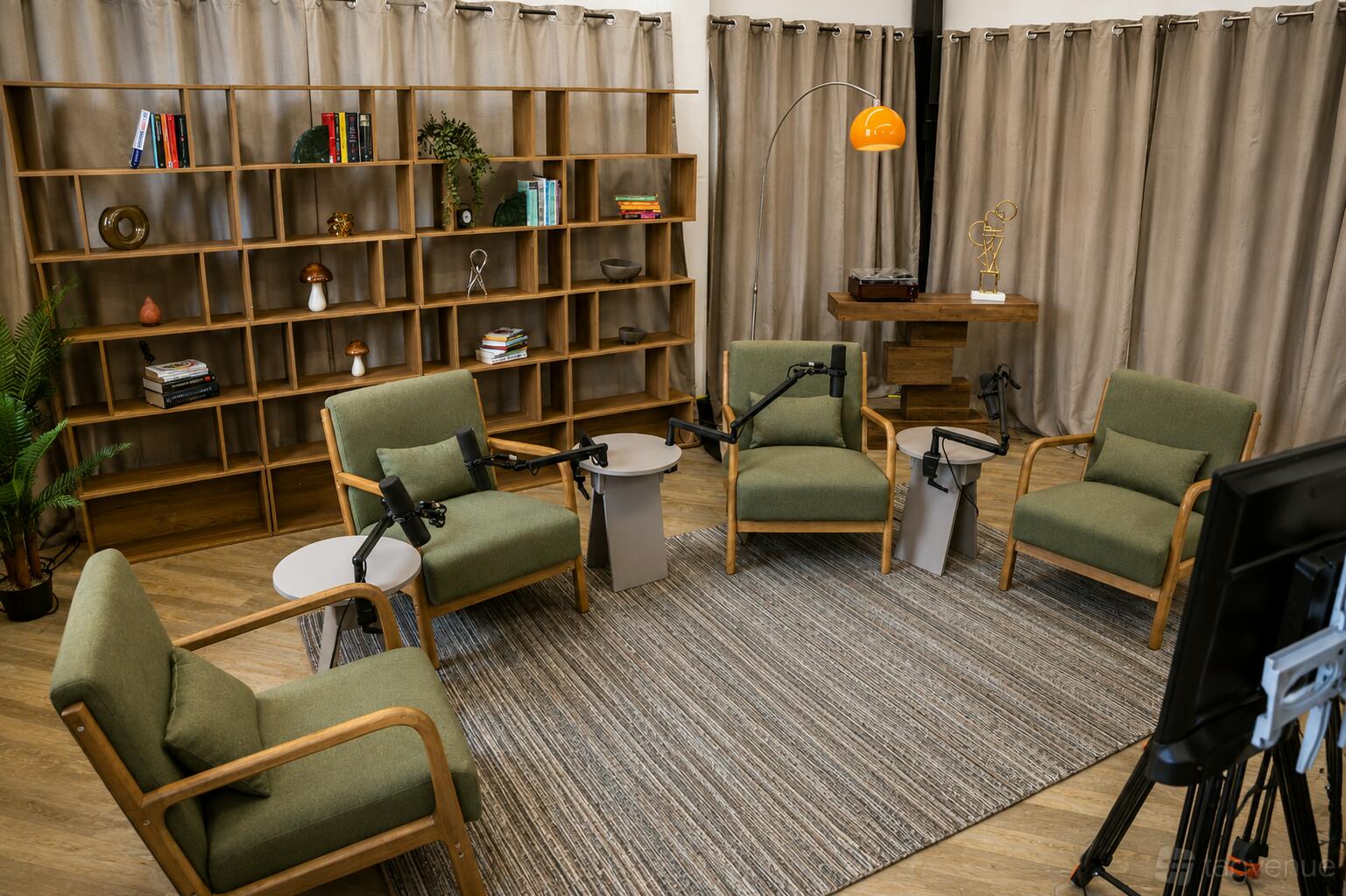 A podcast studio with four green armchairs, microphones, a large bookshelf, and neutral curtains at Podcast Room Camden.