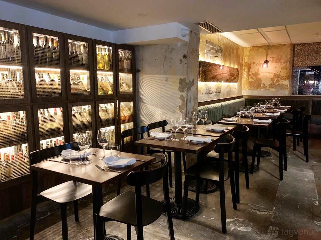 A restaurant basement with a wall of illuminated wine racks, exposed plaster walls, and neatly set dining tables at Figo Ristorante & Bar.