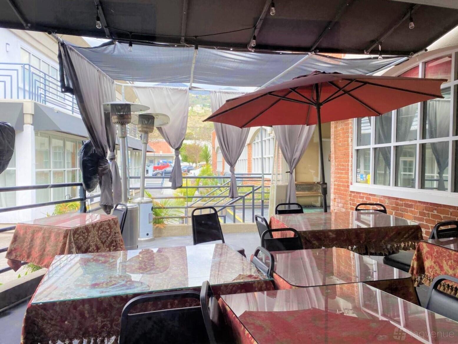 An outdoor patio with covered dining tables, red umbrellas, and patio heaters at Shamshiri Restaurant.