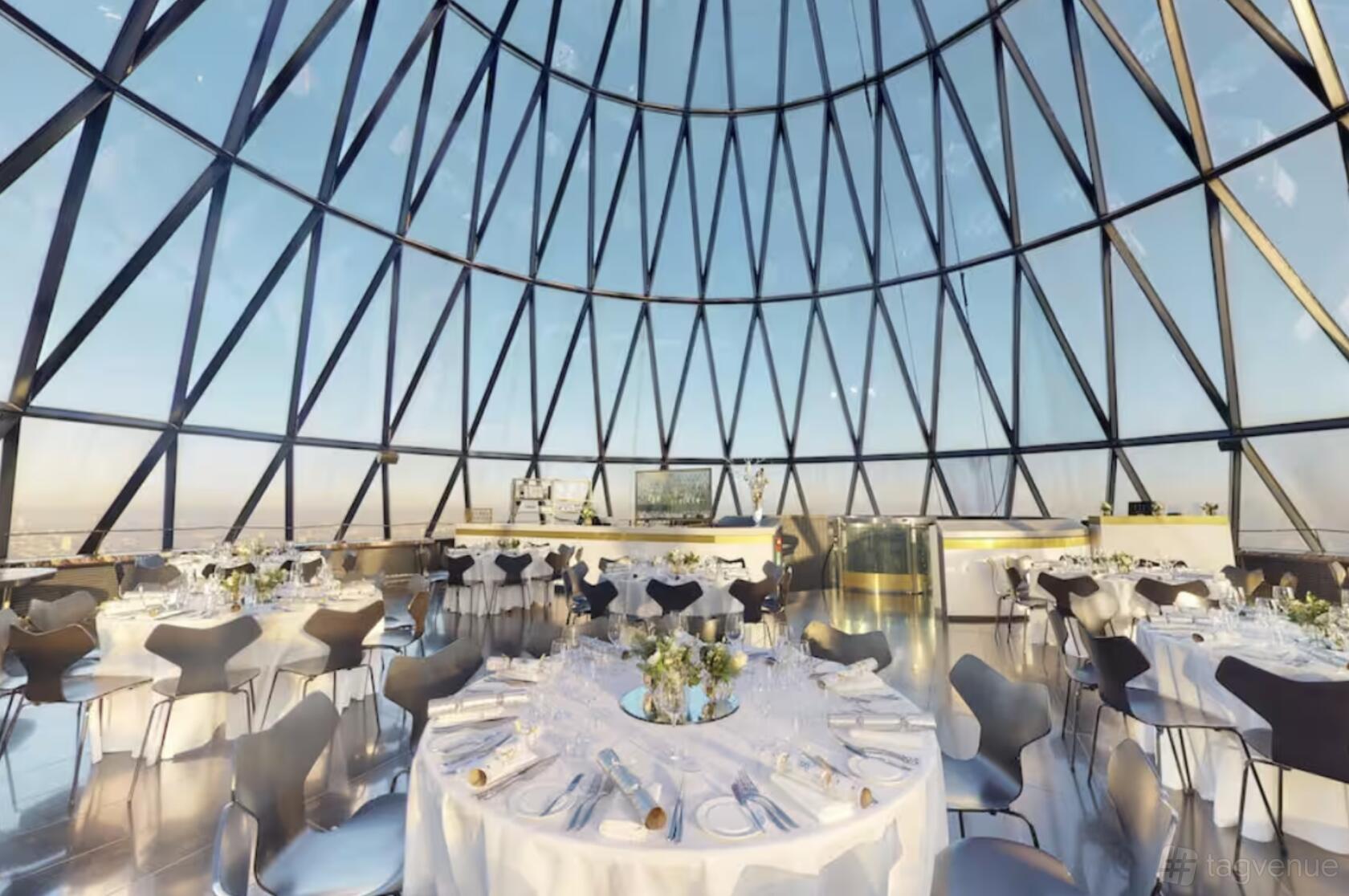 An event space in a restaurant with floor-to-ceiling glass windows, round tables with white linens, and city views at Searcys at The Gherkin.