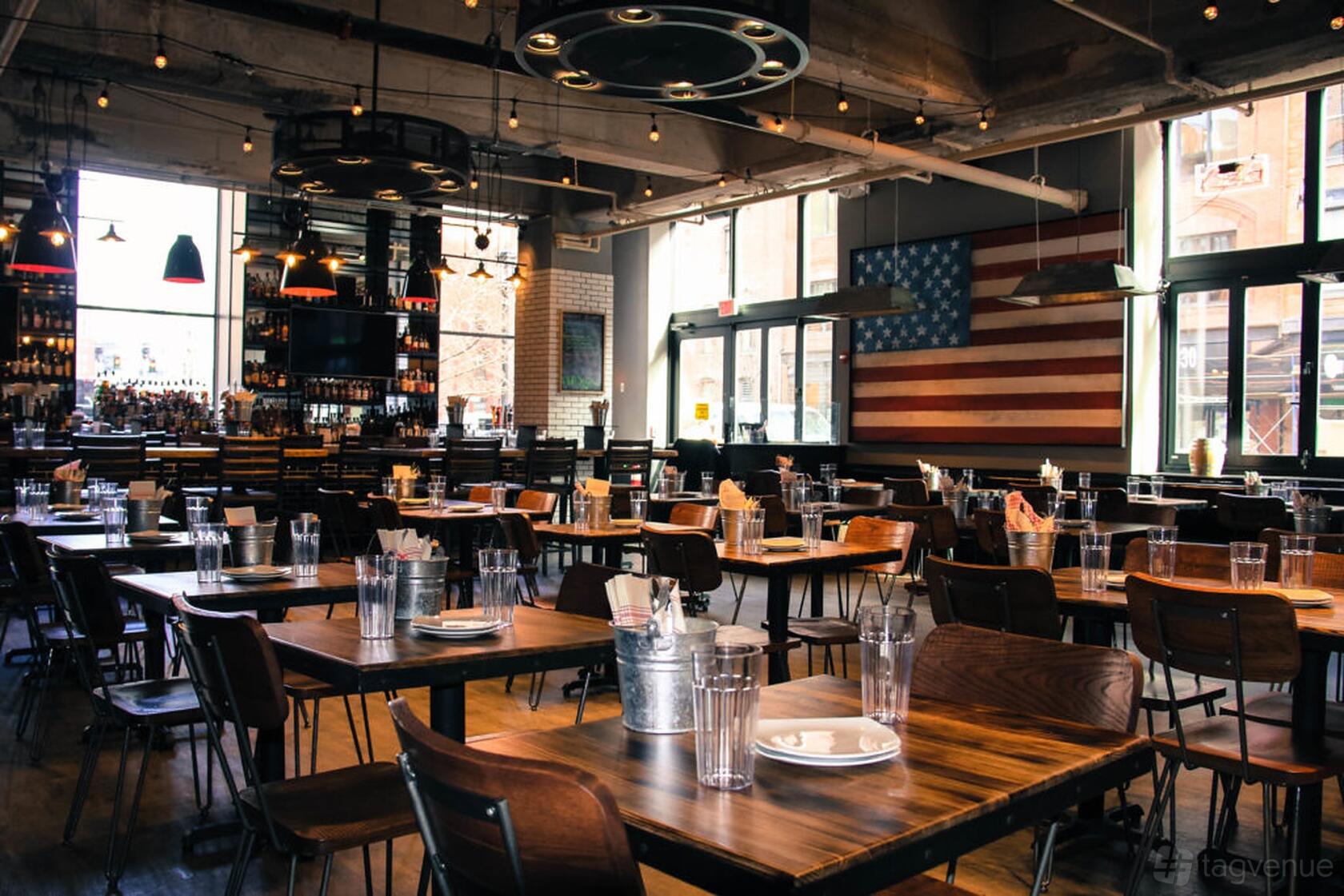 A restaurant with wooden tables, metal chairs, string lights, and an American flag mural at The Smoke Shop BBQ - Seaport.