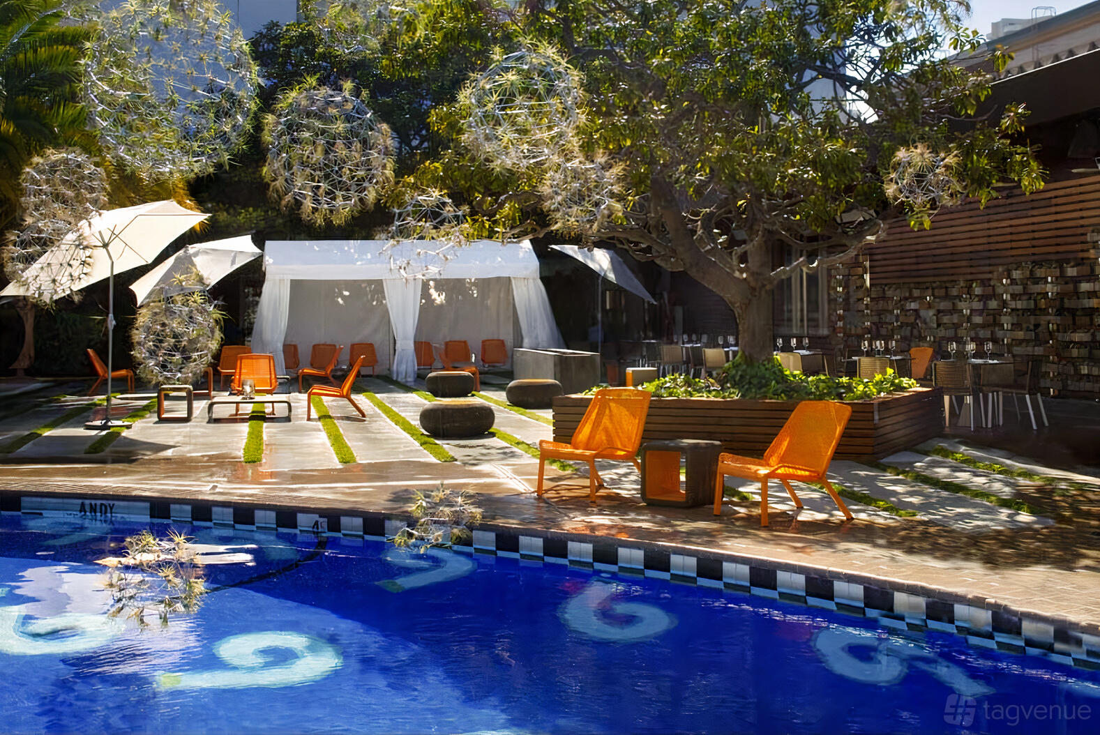 An outdoor hotel space with a pool, orange chairs, large trees, and a white event tent at Chambers at the Phoenix.