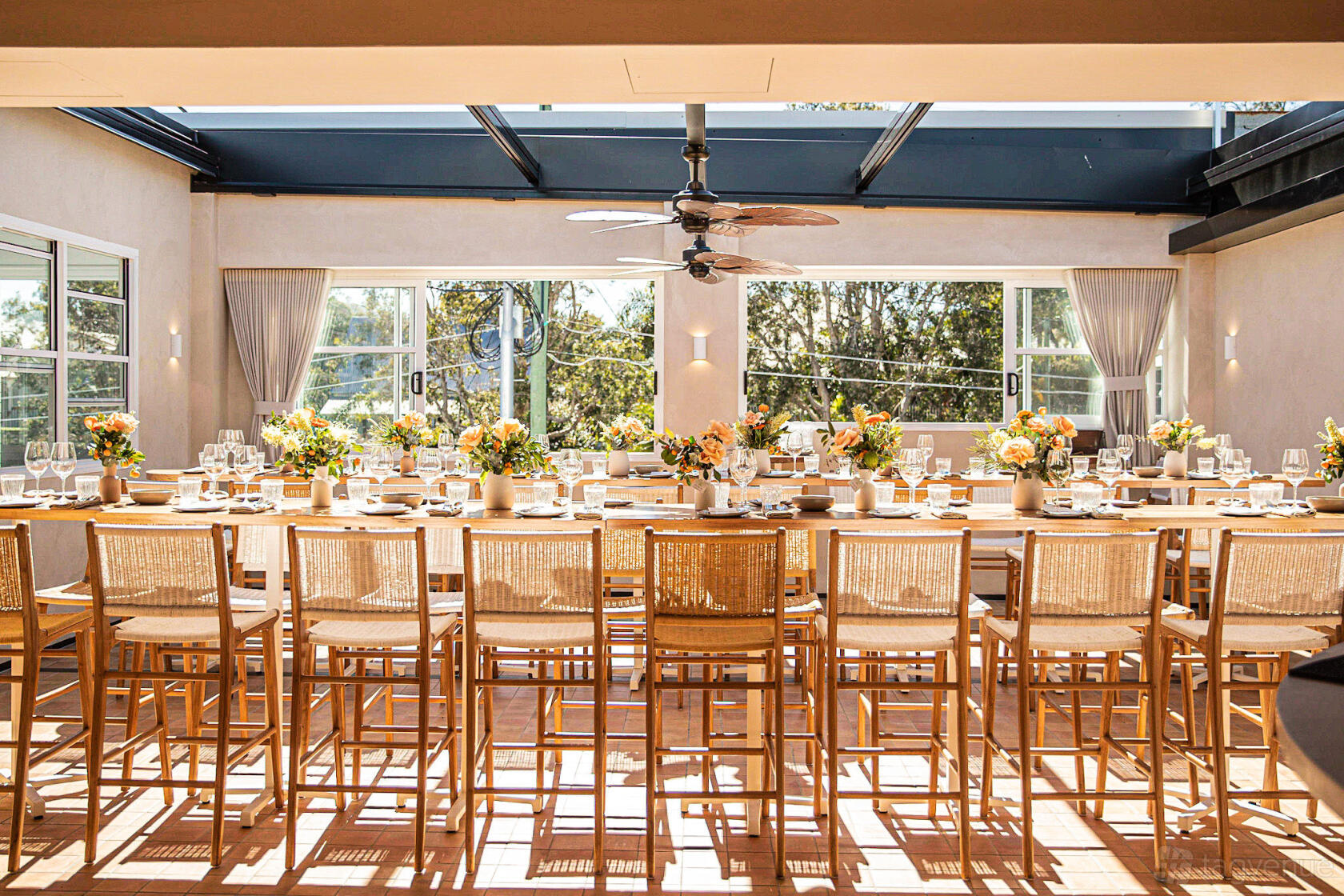 An event space in a restaurant with floor-to-ceiling windows, rattan chairs, and long wooden tables set with floral centerpieces at Melrose Restaurant & Rooftop Bar.