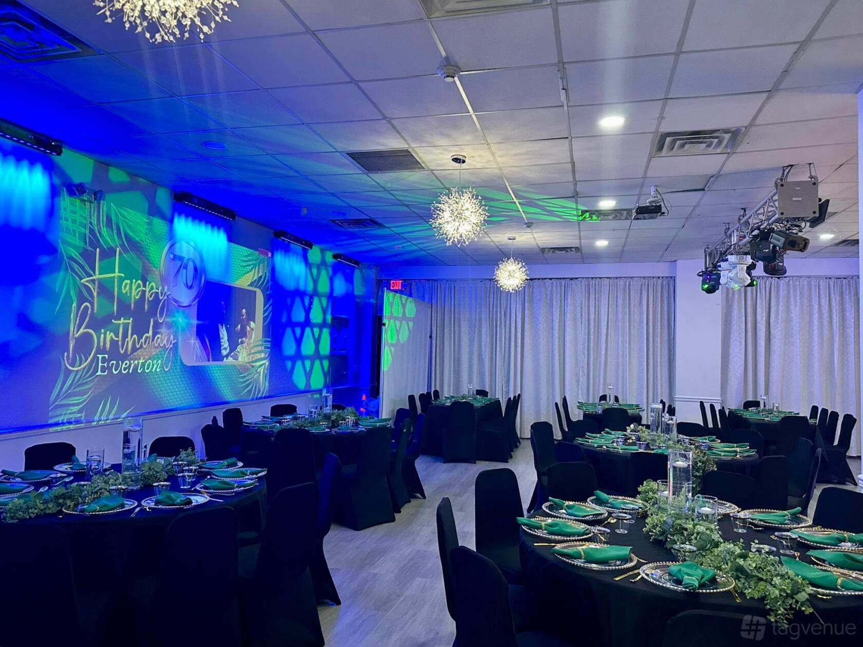 A banquet hall with round tables set for dining, black chairs, blue lighting, and decorative wall projections at Regency Party Hall.