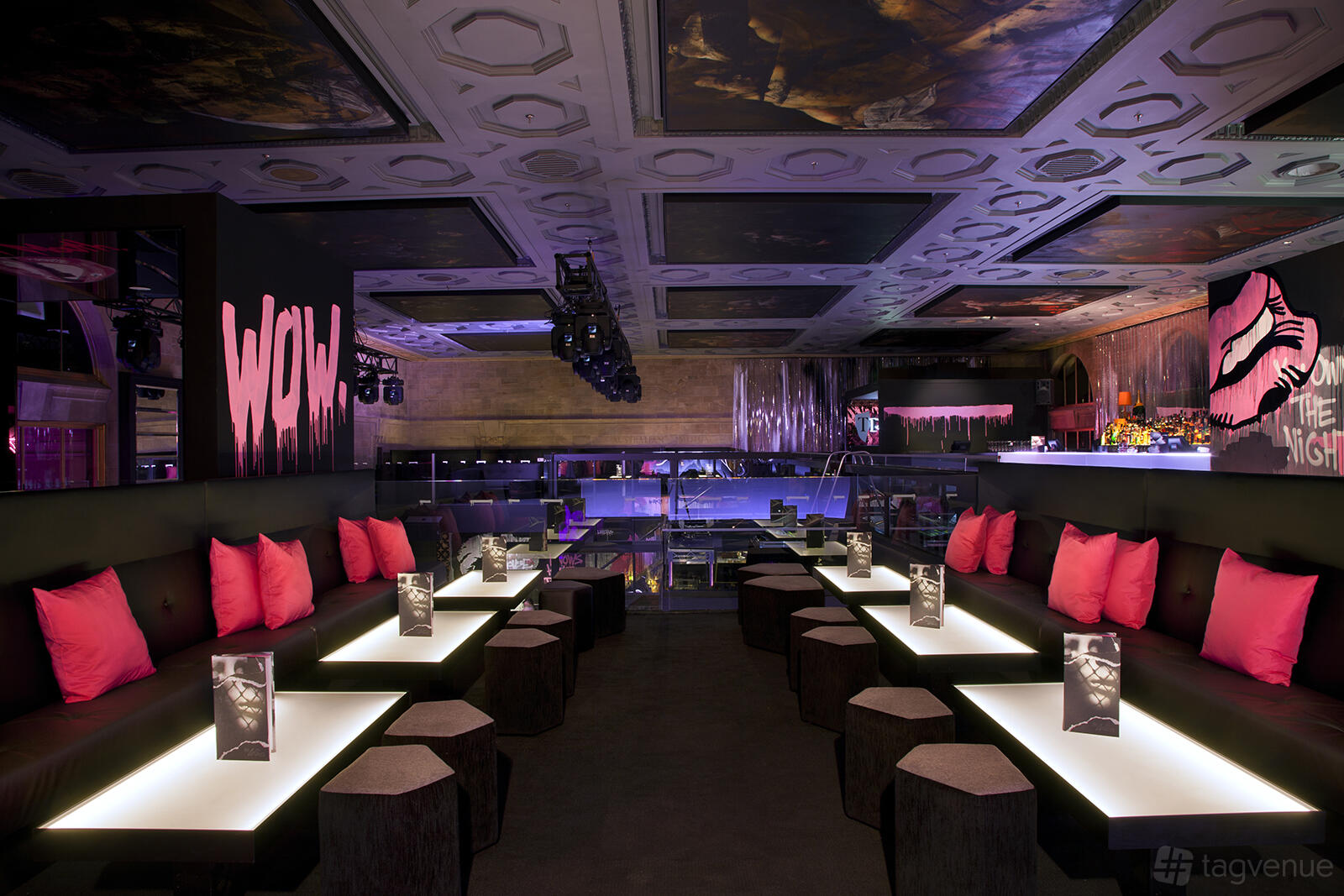 A nightclub lounge with illuminated tables, black seating, and pink accent pillows at Ms Collins.