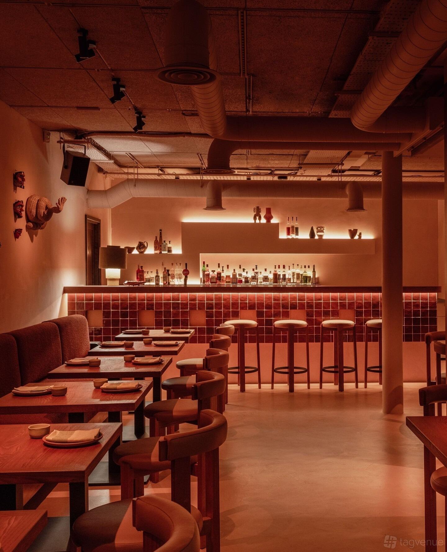A dining room with a tiled bar, backlit shelves with bottles, and wooden tables set for dinner at Fonda.