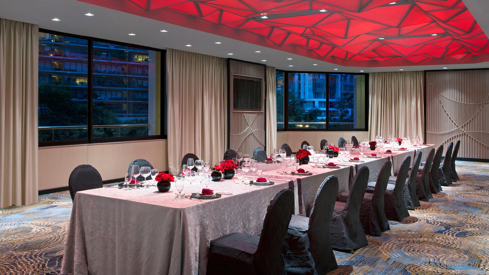 A function room with long banquet tables draped in white linens, red floral centerpieces, and a geometric red ceiling at Four Points by Sheraton.