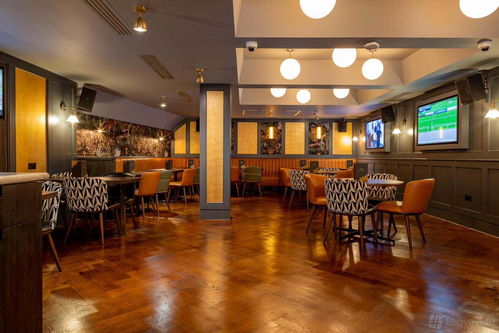 An event space in a pub with round tables, patterned chairs, wall-mounted TVs, and globe pendant lights at Goldwood.