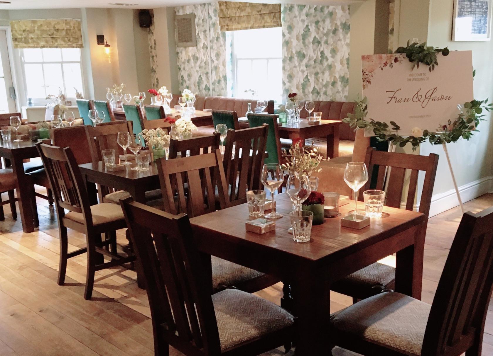 A dining room in a pub with wooden tables set with glassware, floral centerpieces, and large windows at Inn on The Lake.