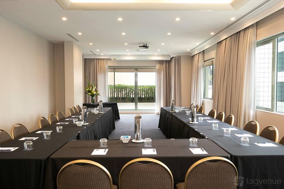 A hotel meeting room with U-shaped tables, black tablecloths, notepads, water glasses, and large windows at The Sebel Sydney Chatswood.
