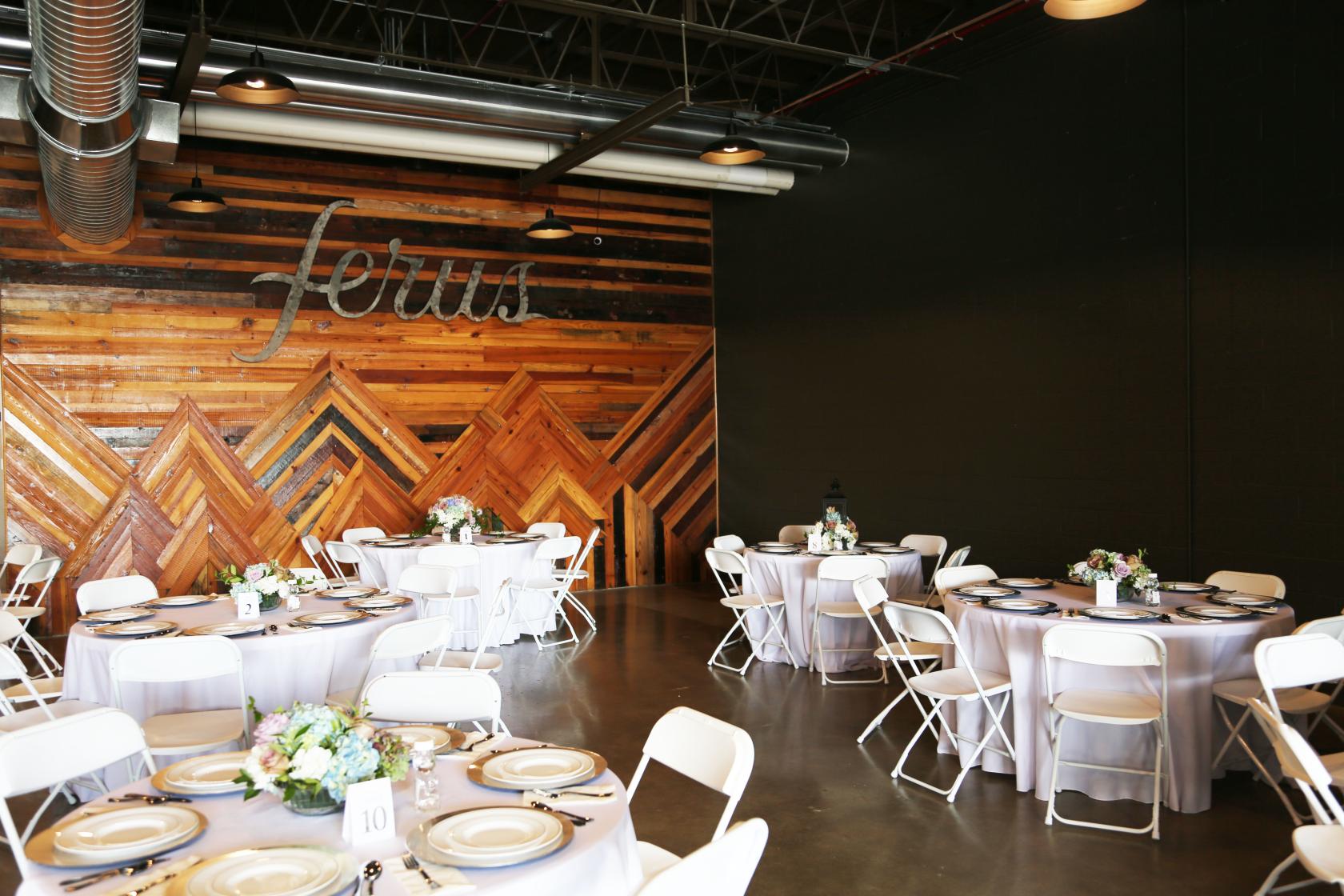 An event venue with wooden accent walls, exposed ductwork, and round tables with white linens at Ferus Artisan Ales.