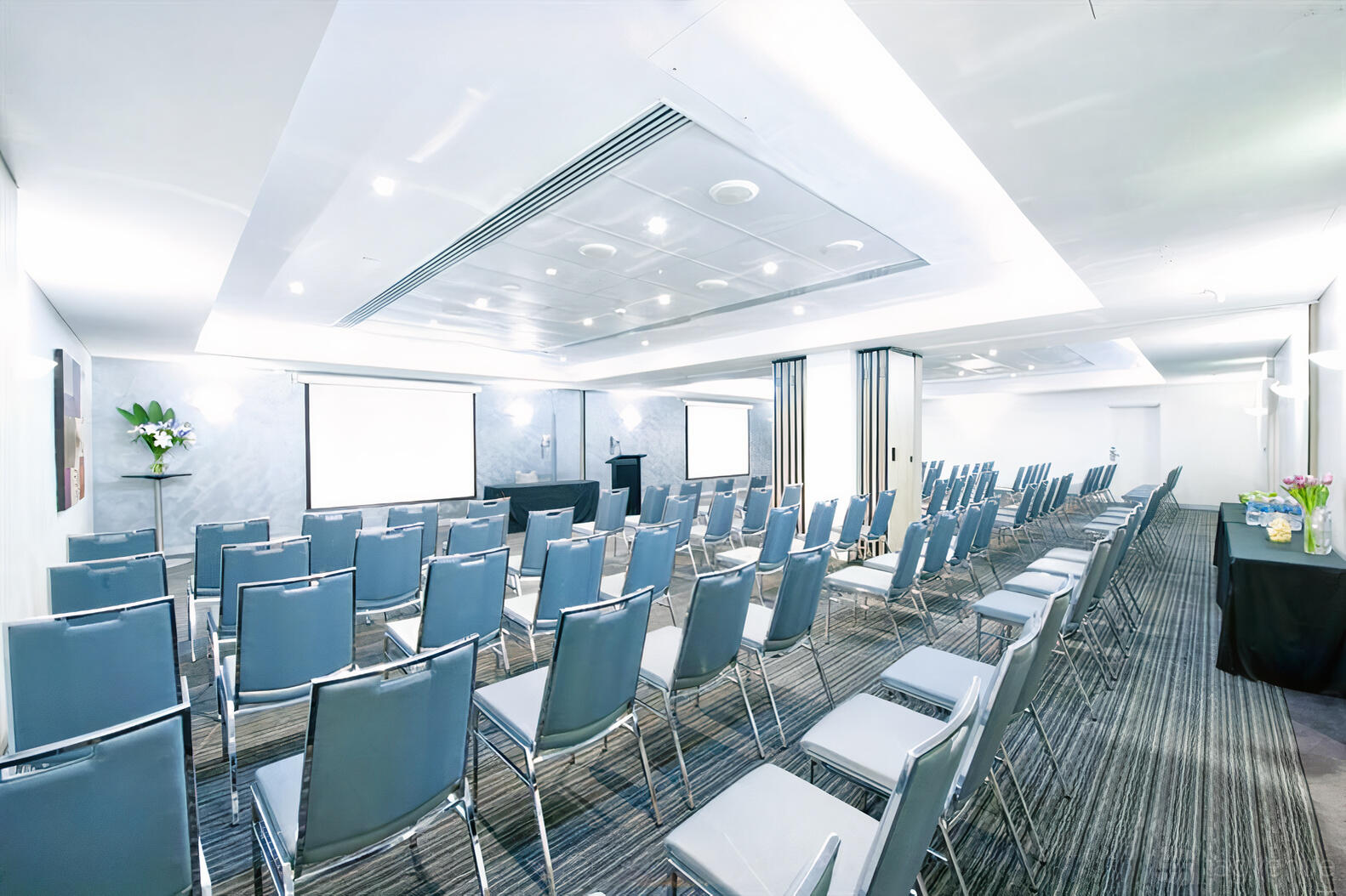 A hotel meeting room with rows of grey chairs, ceiling-mounted projectors, and two projection screens at Mantra Chatswood.