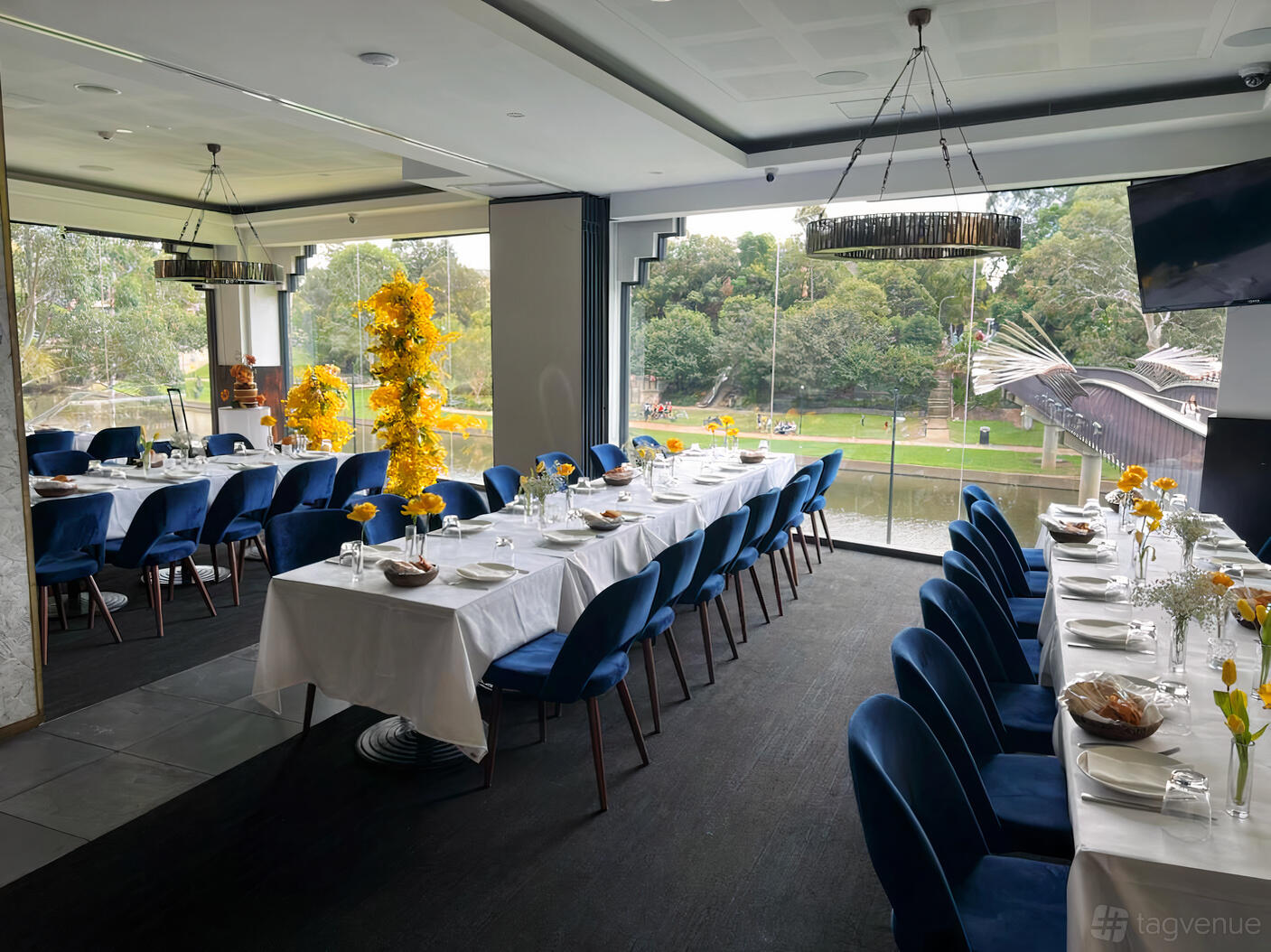A restaurant dining room with floor-to-ceiling windows, river views, blue chairs, and white-clothed tables at Sahra By The River.