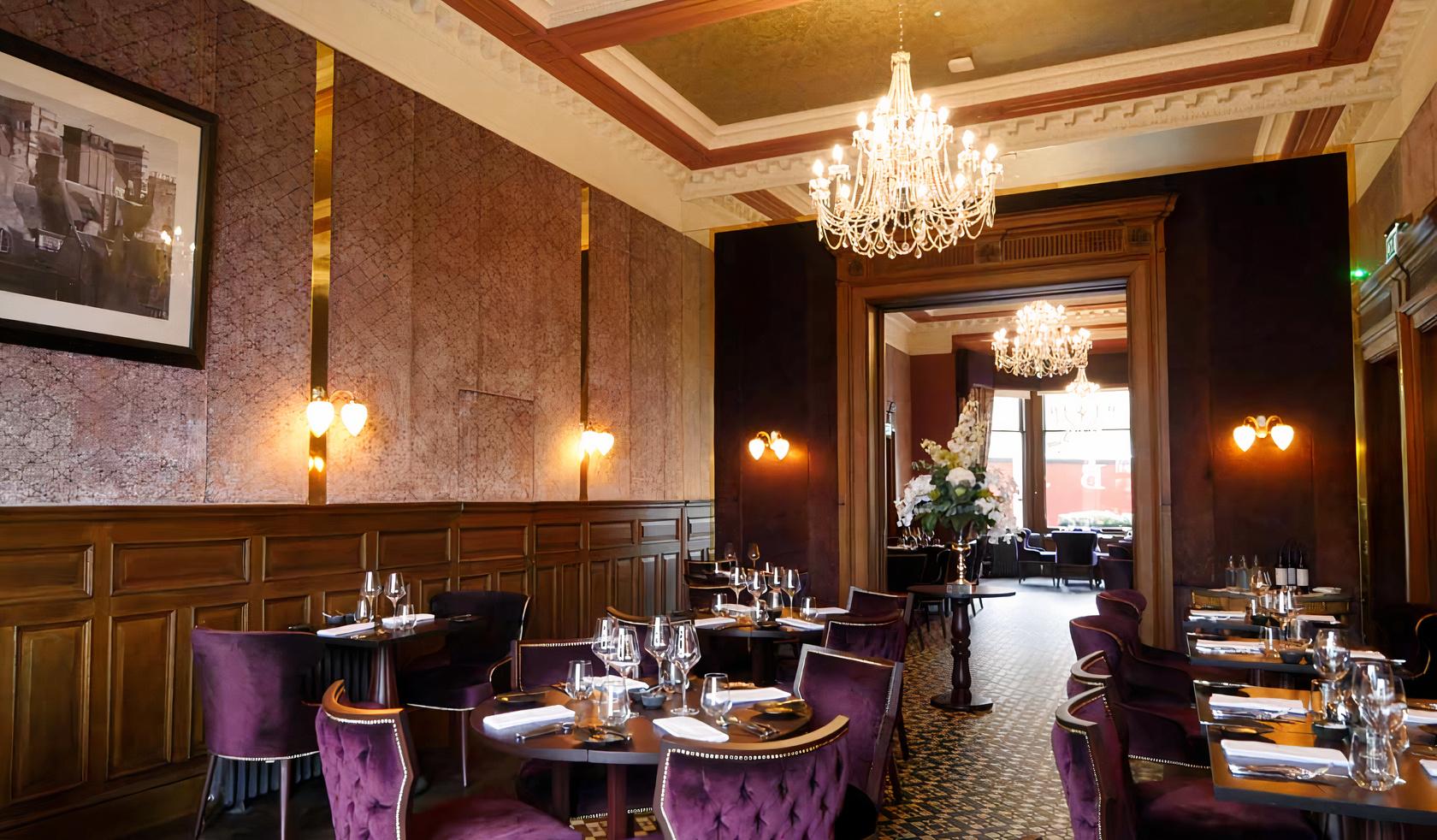A restaurant with velvet dining chairs, chandeliers, wood paneling, and large windows at The Bonham Hotel.