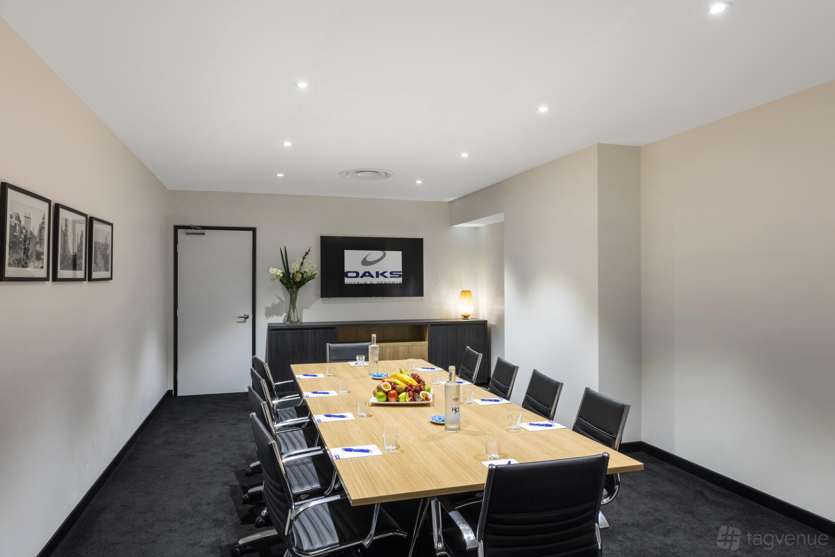 A hotel meeting room with a long wooden table, black chairs, fruit platter, and wall-mounted screen at Oaks Embassy.