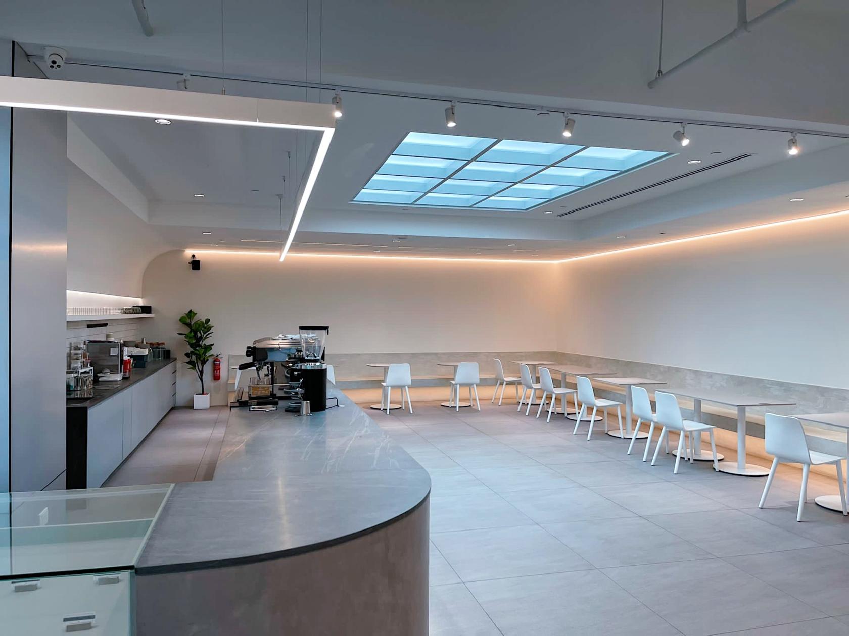 A cafe with minimalist decor, skylight ceiling, light grey flooring, and white tables and chairs at SYIP (Luzerne).