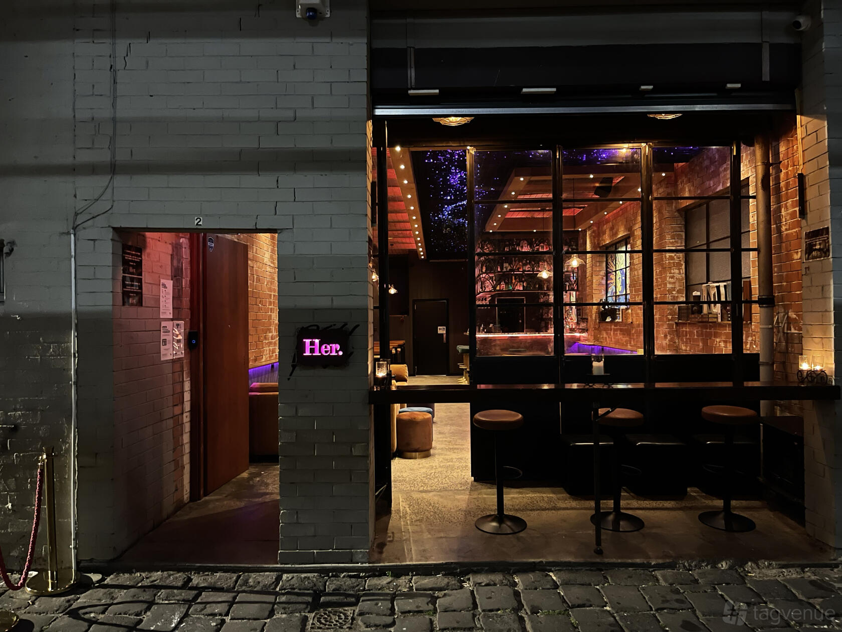A bar with exposed brick walls, high stools, and purple accent lighting at Her.