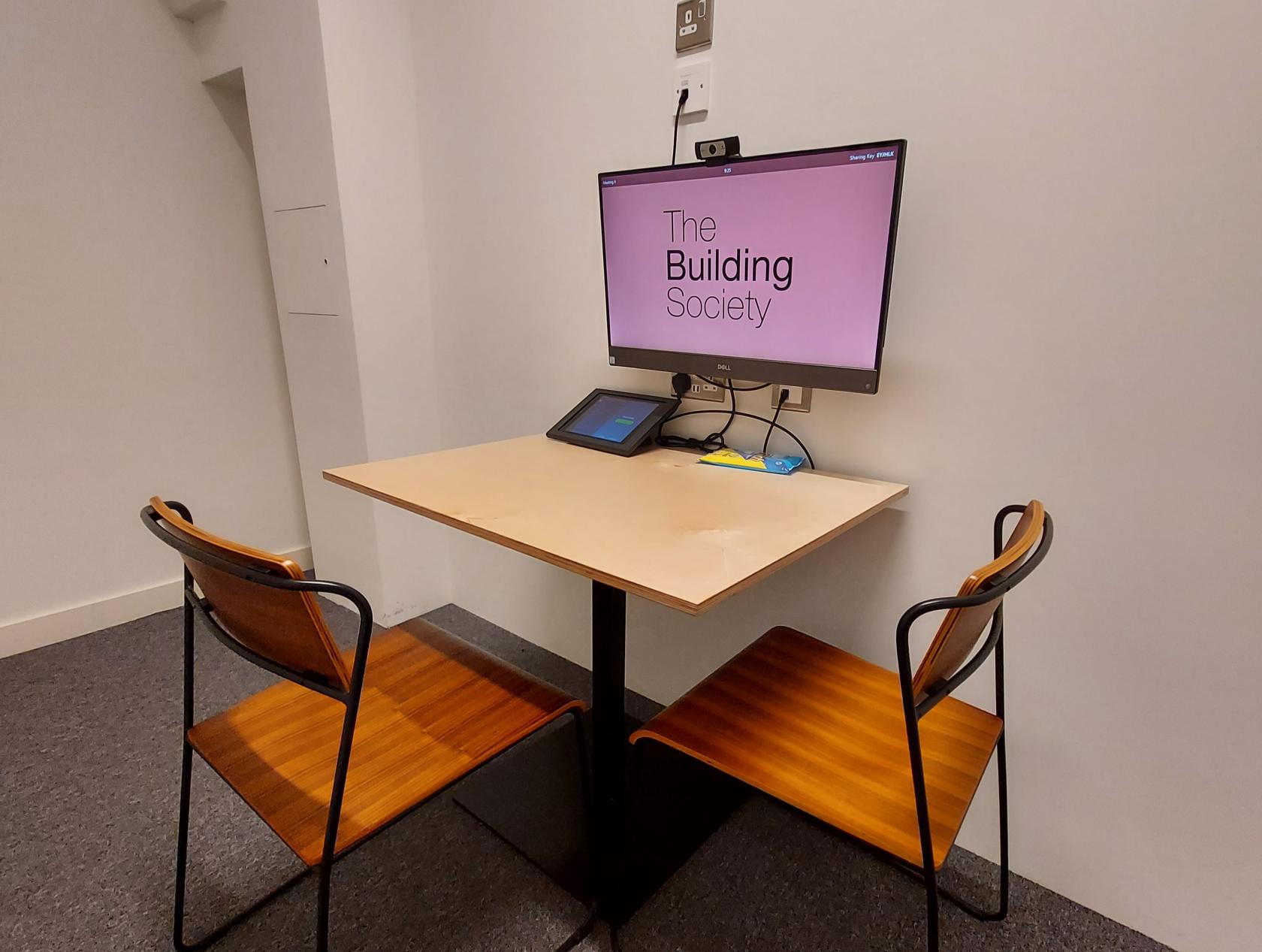 A meeting room with a small table, two wooden chairs, and a wall-mounted screen at The Society Building - Fitzrovia.