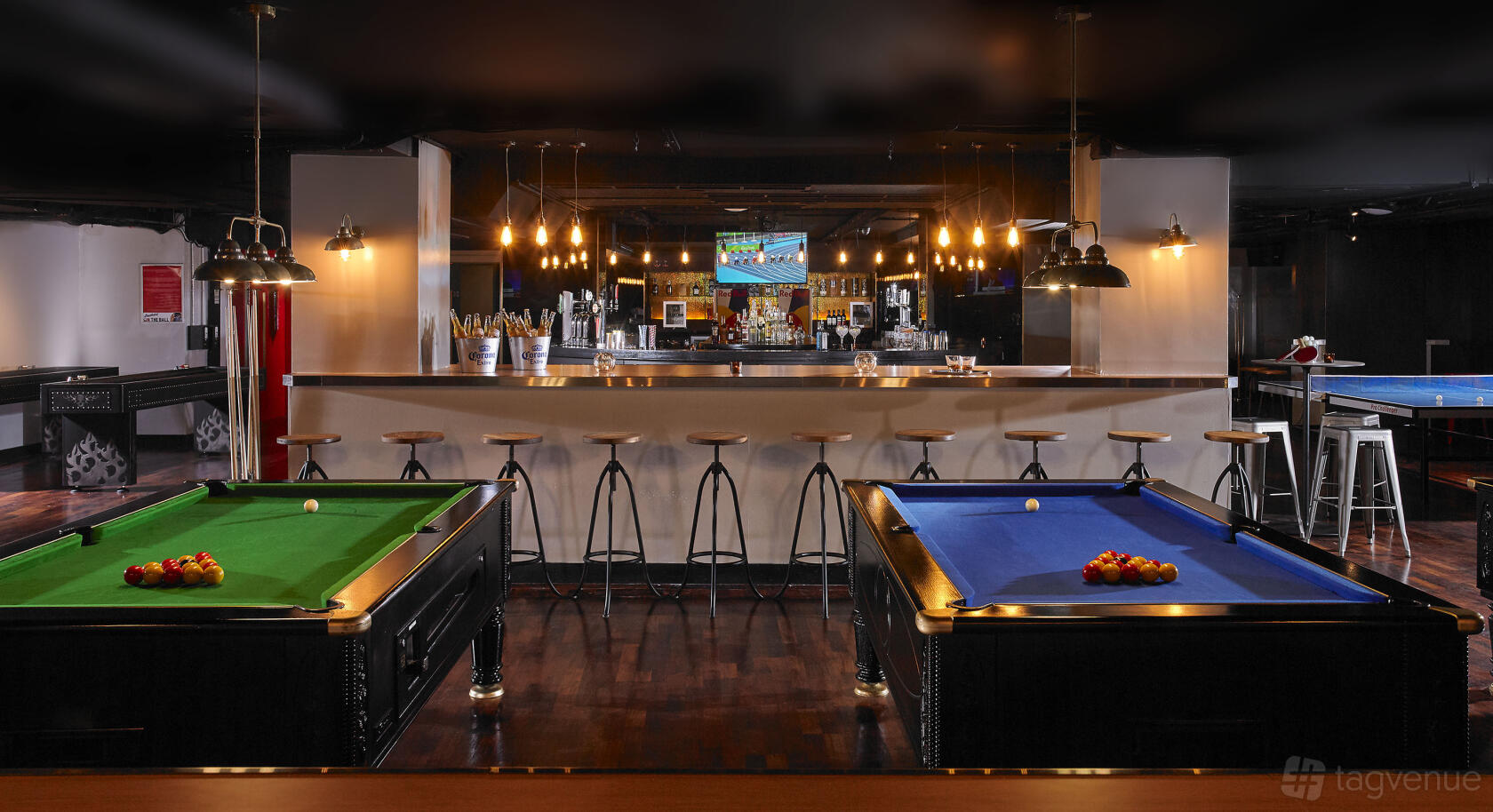 A bar with pool tables, hanging pendant lights, bar stools, and a large TV screen at Buskers On The Ball.