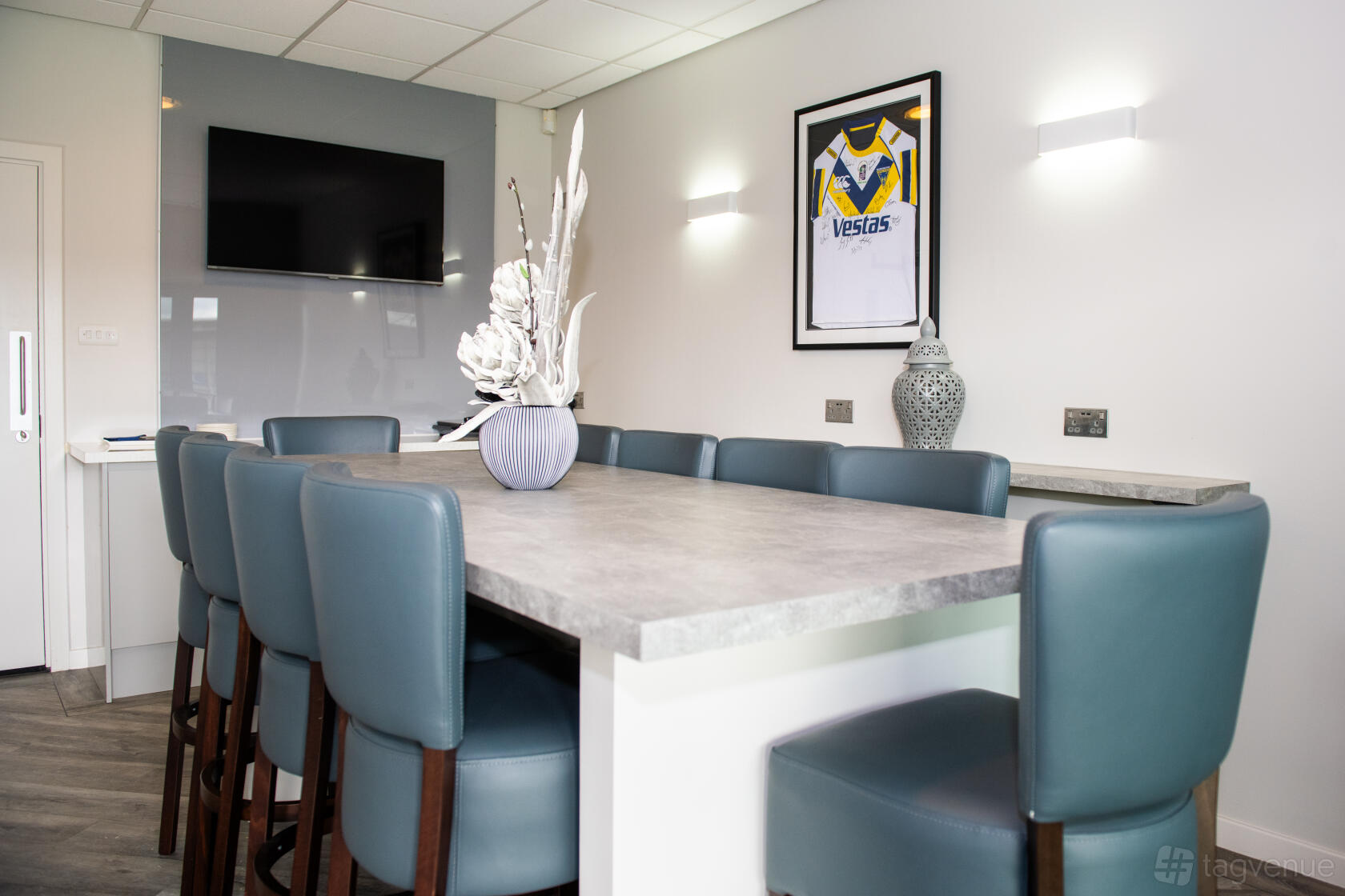 An executive suite with a long stone-top table, blue leather chairs, and framed rugby jersey at The Halliwell Jones Stadium.