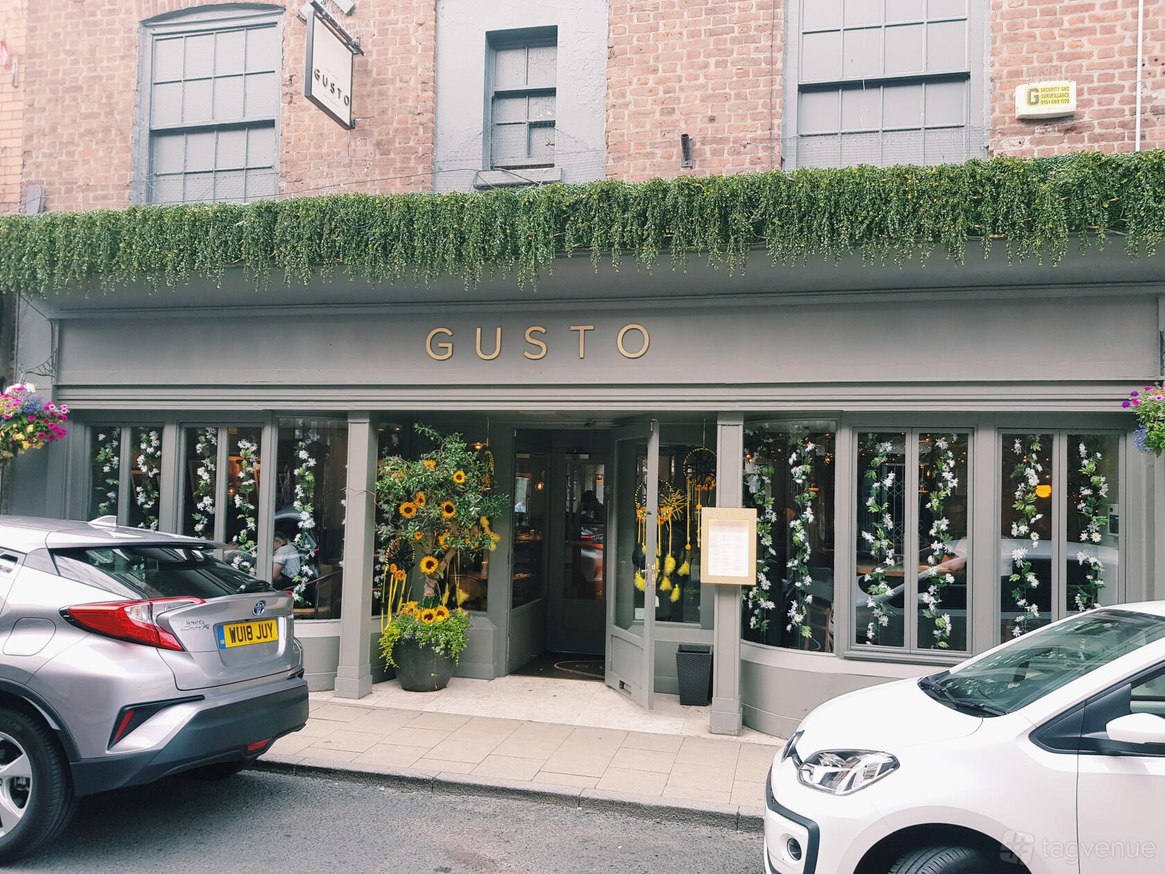 A restaurant entrance with grey panelled windows, hanging greenery, and flower arrangements at Gusto Restaurant and Bar Knutsford.