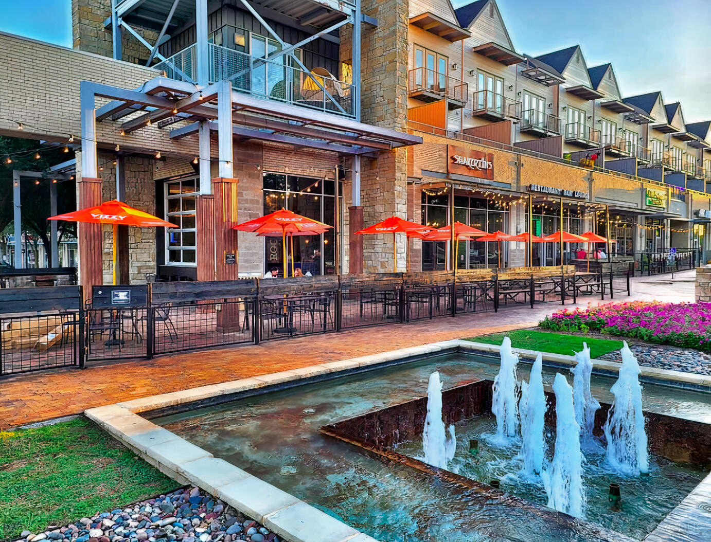 A pub with outdoor patio seating, red umbrellas, and a small fountain in front at Shakertins -The Colony.