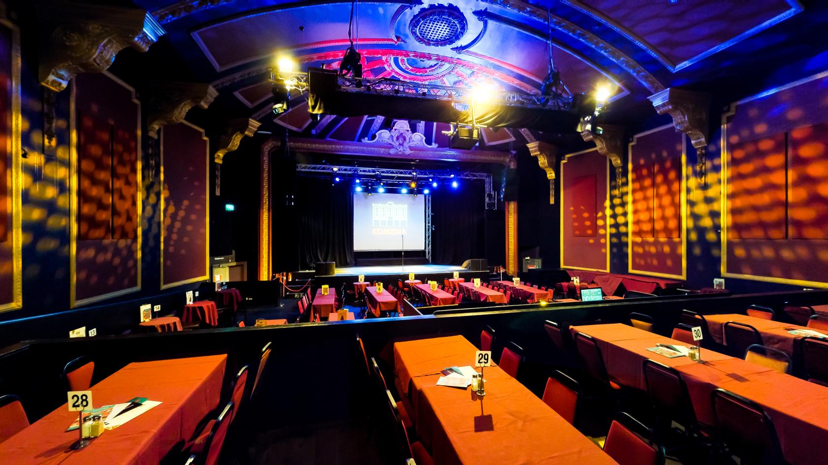 An event venue with rows of rectangular tables facing a stage with theater lighting at Komedia.