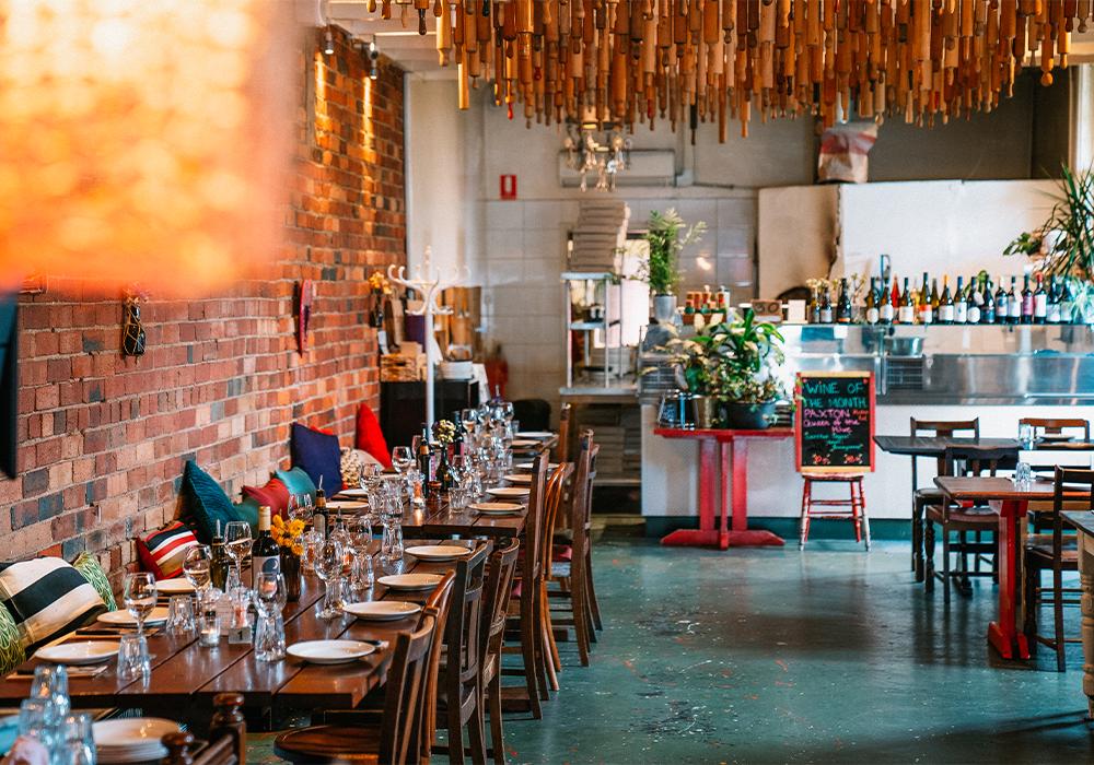 A restaurant with exposed brick walls, long wooden tables set for dining, and a ceiling of hanging wooden dowels at Farro Thornbury.