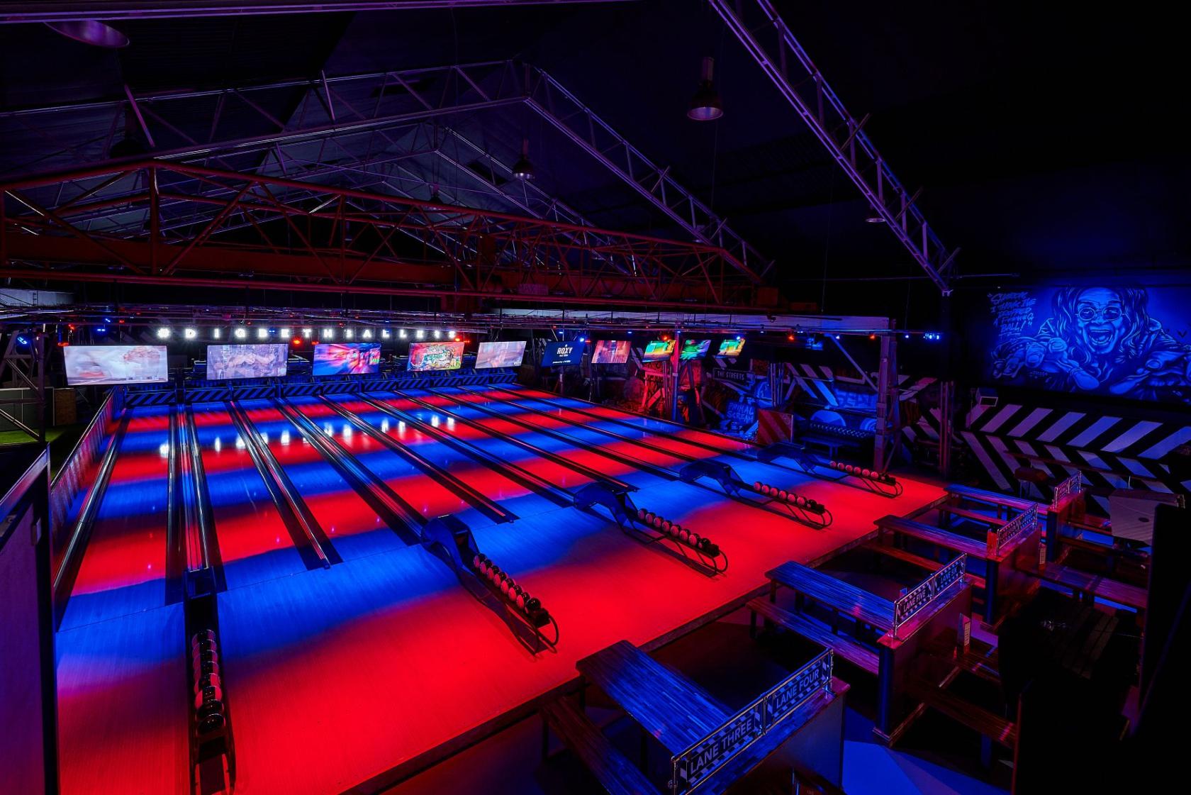 A bowling alley with neon-lit lanes, multiple overhead screens, and wooden seating at Roxy Lanes Birmingham Digbeth.