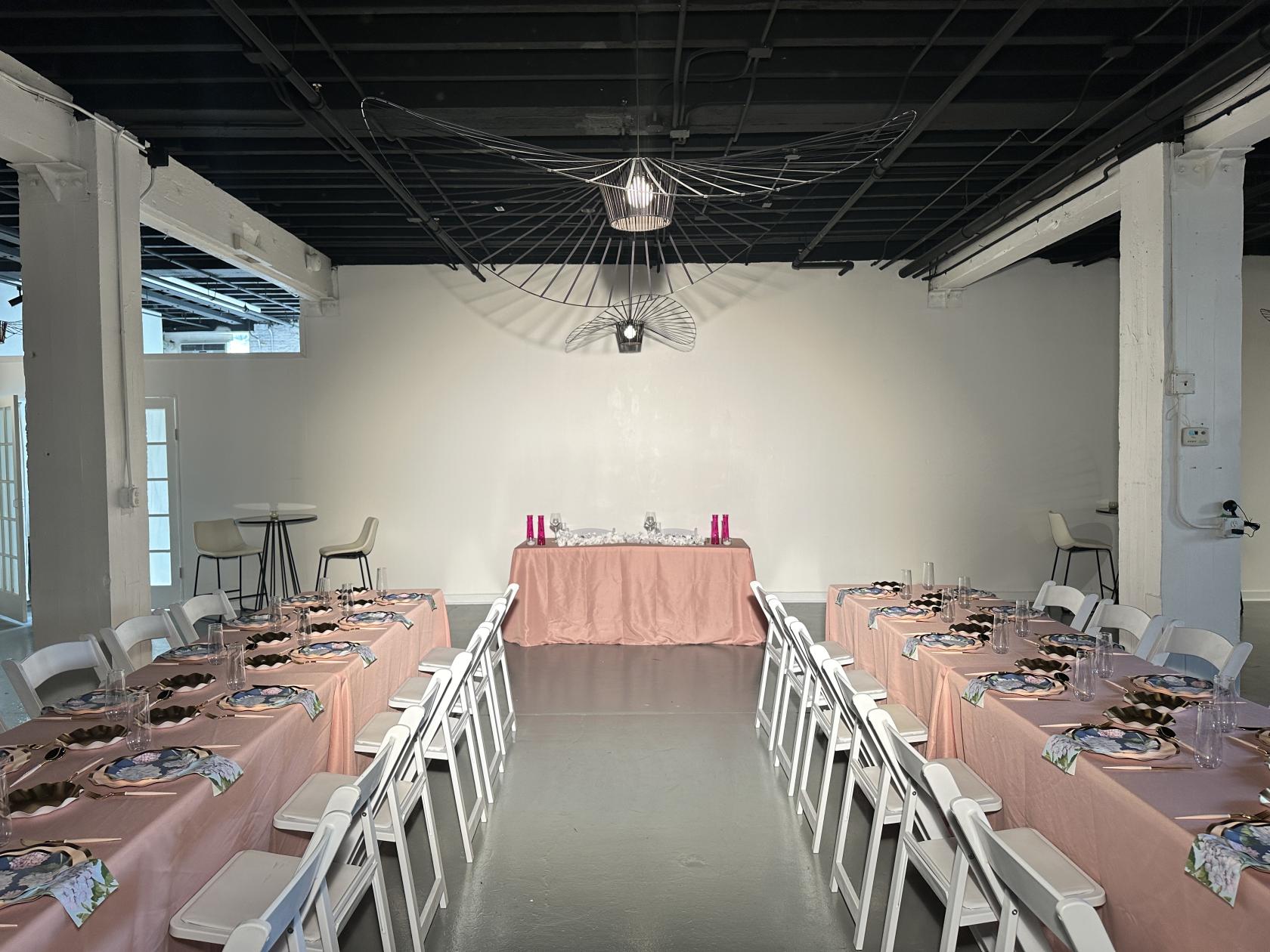 An event venue with white walls, exposed black ceiling, and two long banquet tables with pink linens at The Fulton West.