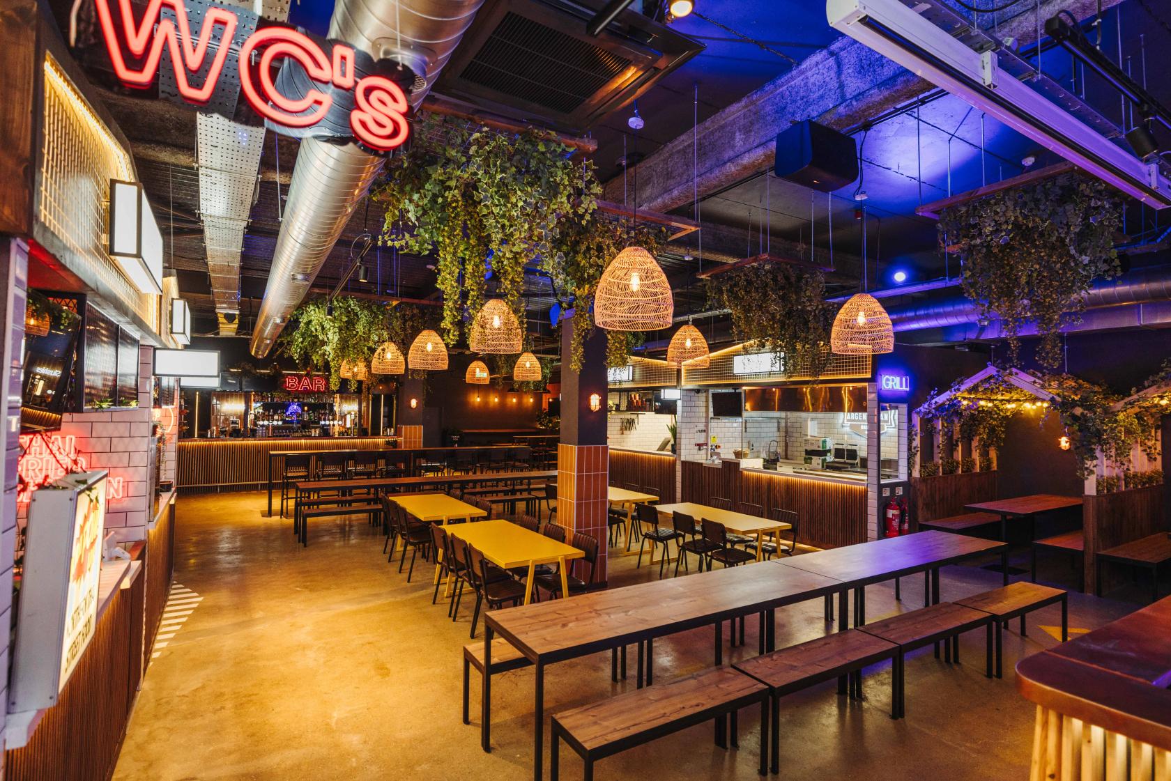 A restaurant with long wooden tables, hanging basket lights, and greenery accents at Market Place Peckham.
