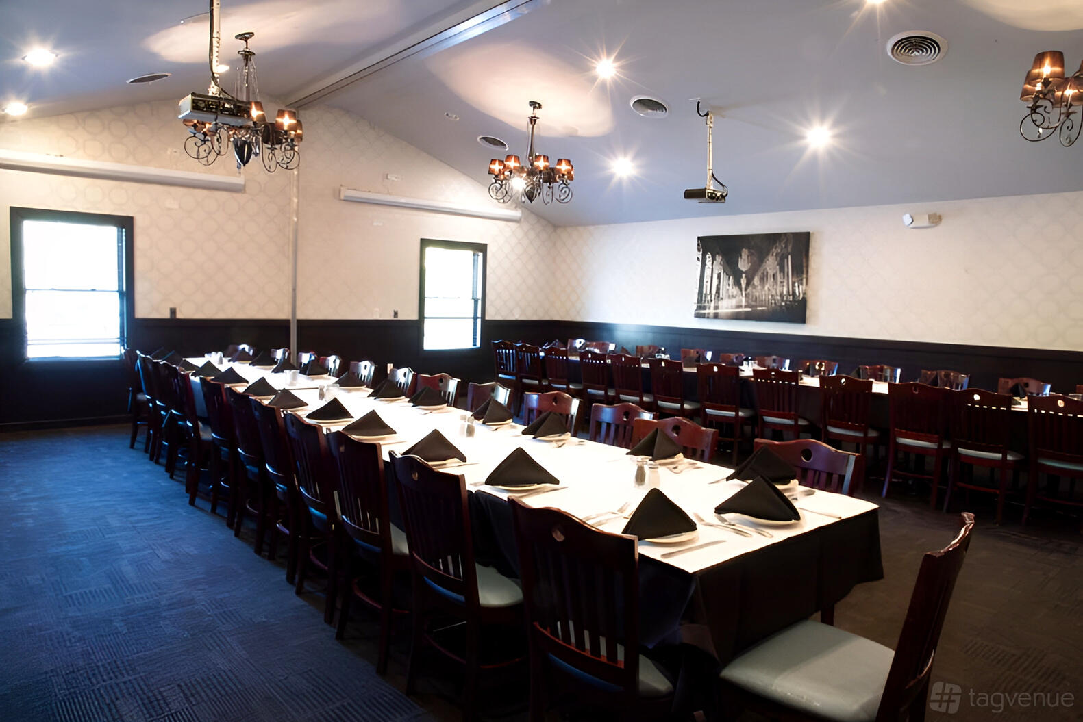 An event space in a restaurant with long banquet tables, dark wooden chairs, chandeliers, and white tablecloths at Gino’s Prime and Surf.