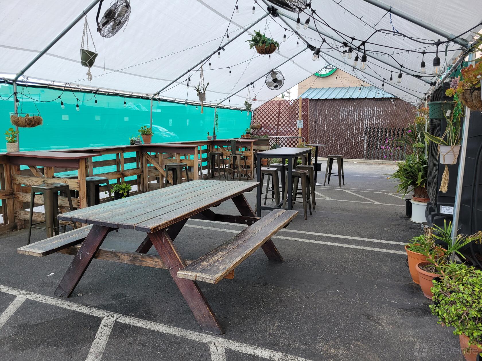 An outdoor brewery area with picnic tables, high-top stools, hanging plants, and string lights at Hand-Brewed Beer.