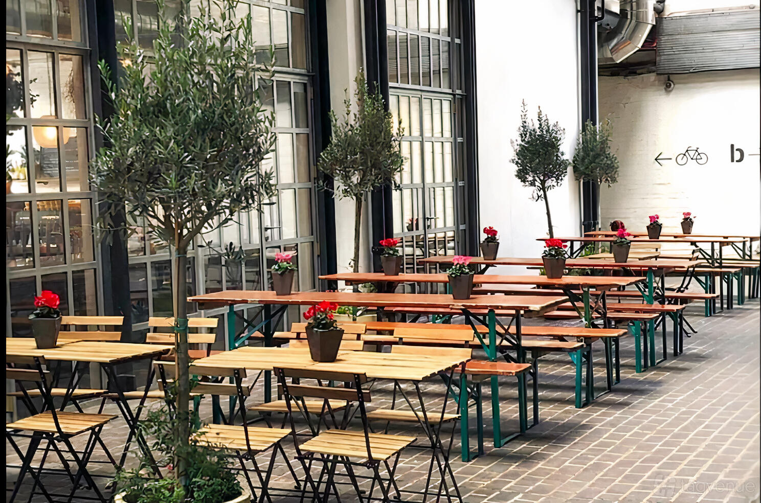 An outdoor restaurant patio with wooden tables, benches, potted flowers, and trees at Fare Restaurant + Bar.
