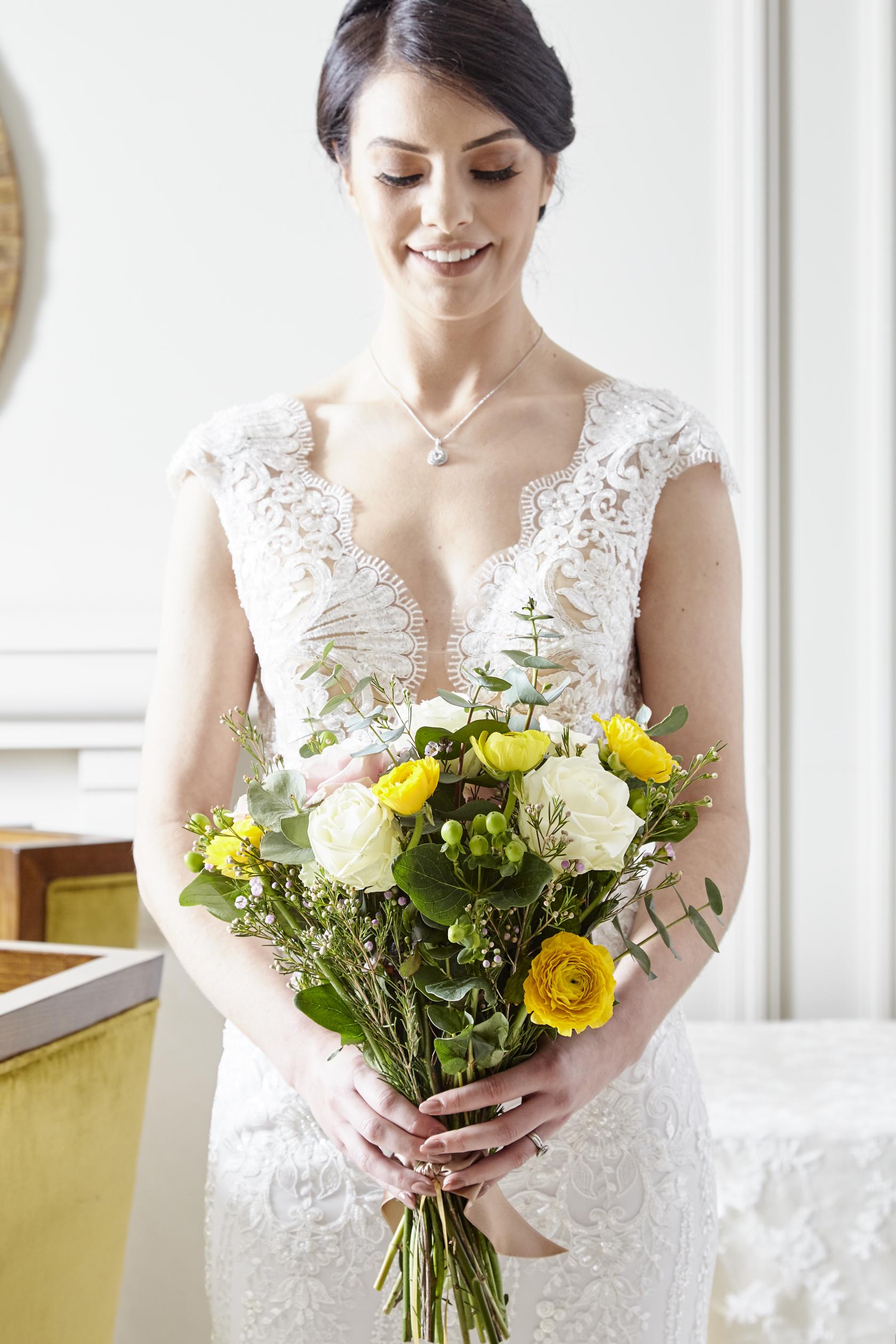 A person in a lace wedding dress holding a bouquet of white and yellow flowers at The Malone Hotel.