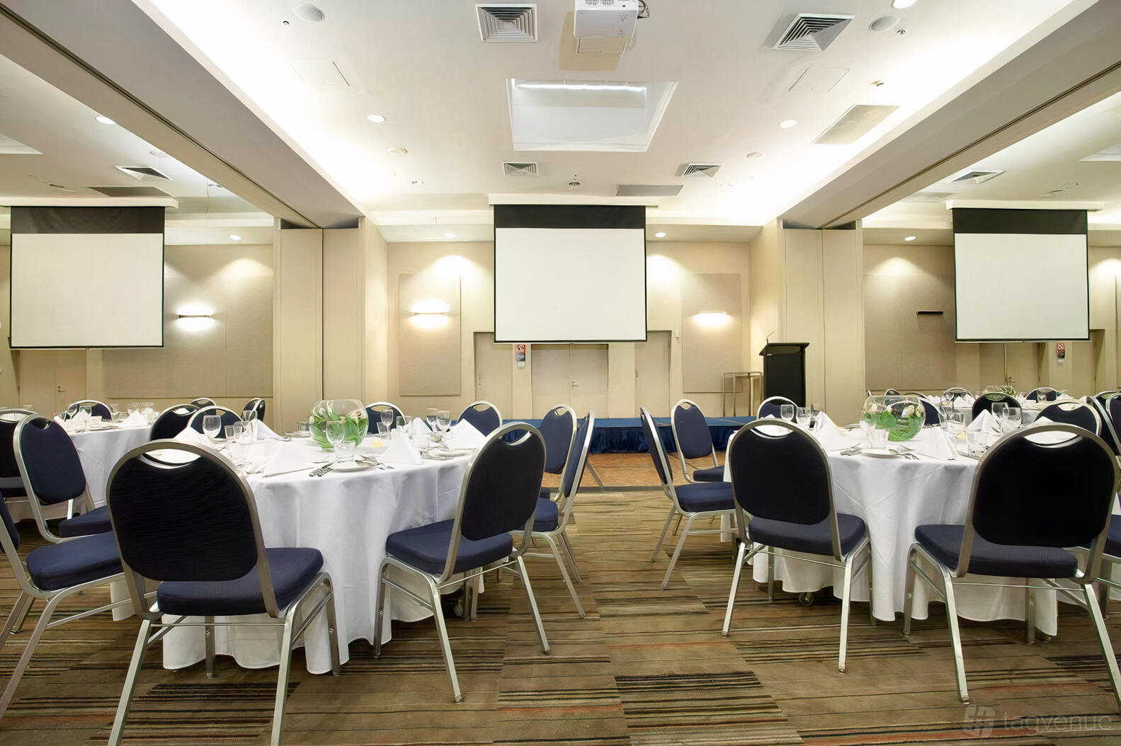 A meeting room with round tables set with white linens, black chairs, and dual projector screens at Novotel Sydney Olympic Park Hotel.