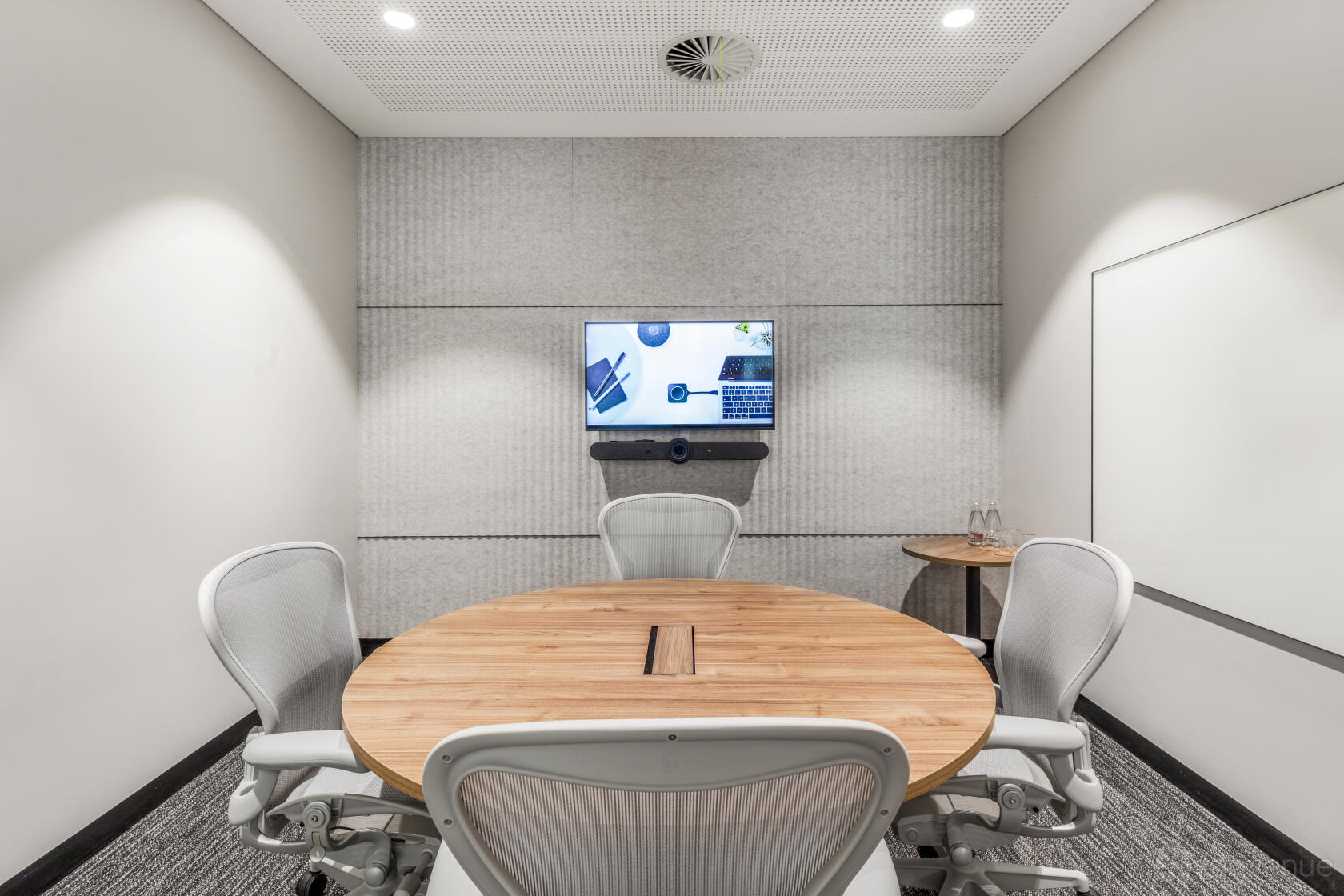 A meeting room with a round wooden table, three ergonomic chairs, wall-mounted screen, and whiteboard at Collins Place.