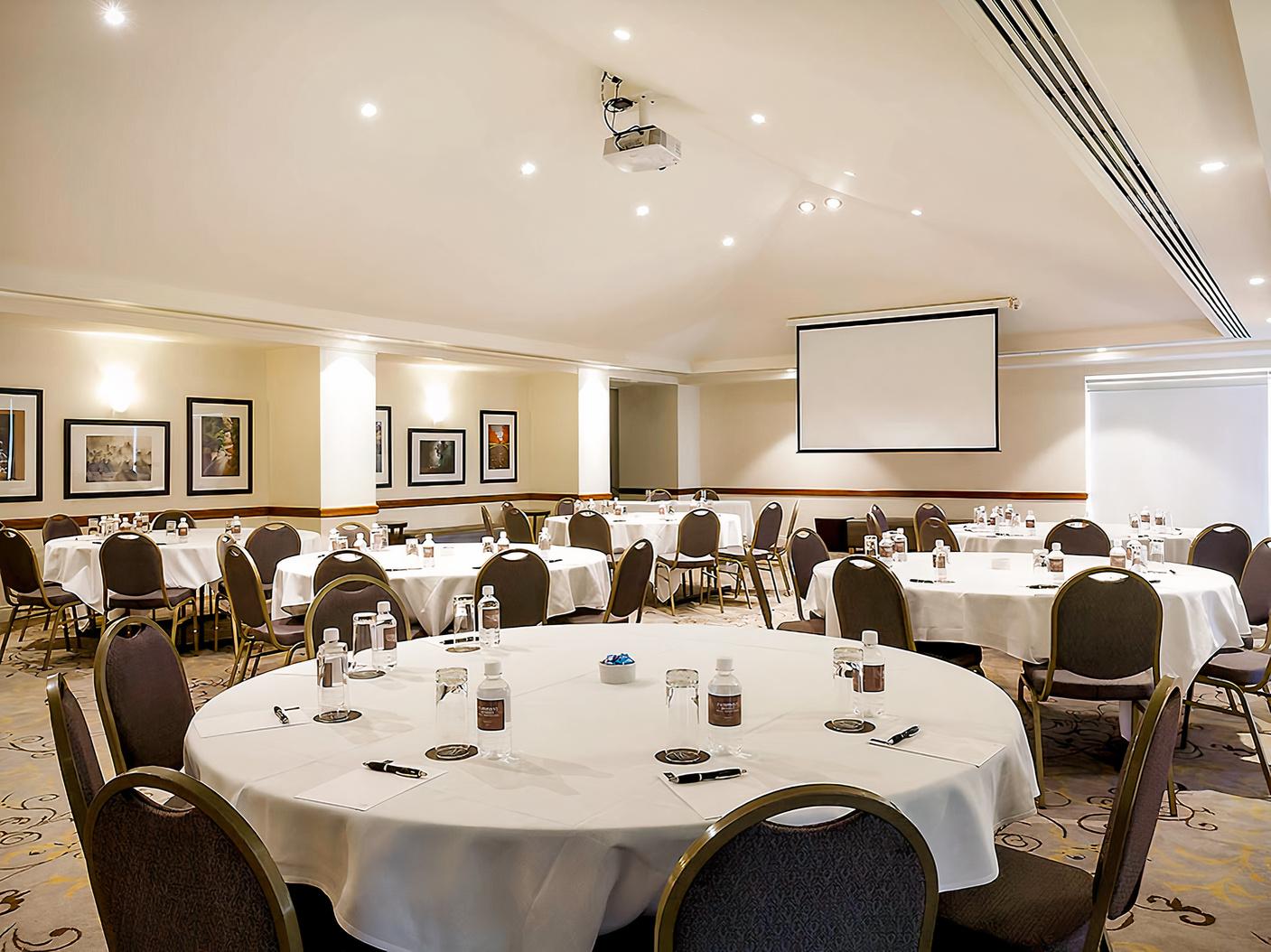An event space in a hotel with round tables, chairs, water glasses, notepads, and a ceiling projector at Fairmont Resort Blue Mountains.