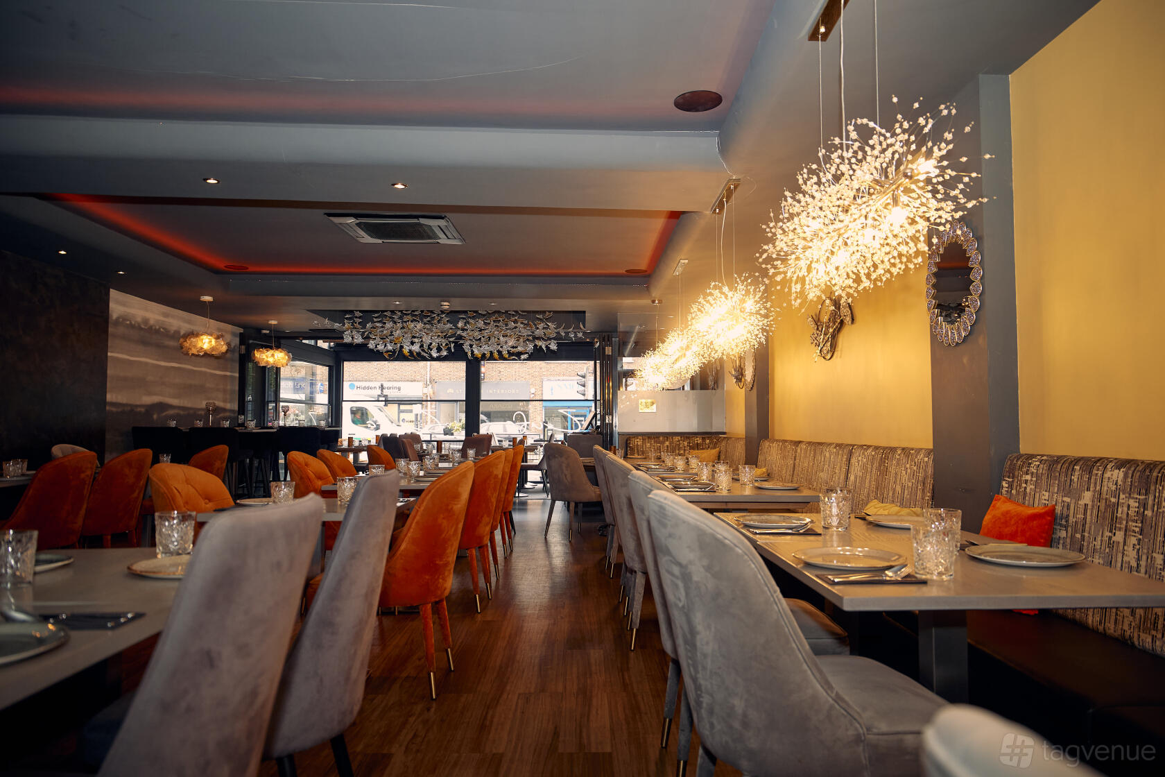 A restaurant with long tables, orange and beige upholstered chairs, decorative lighting, and wooden floors at Zufa Hatch End.