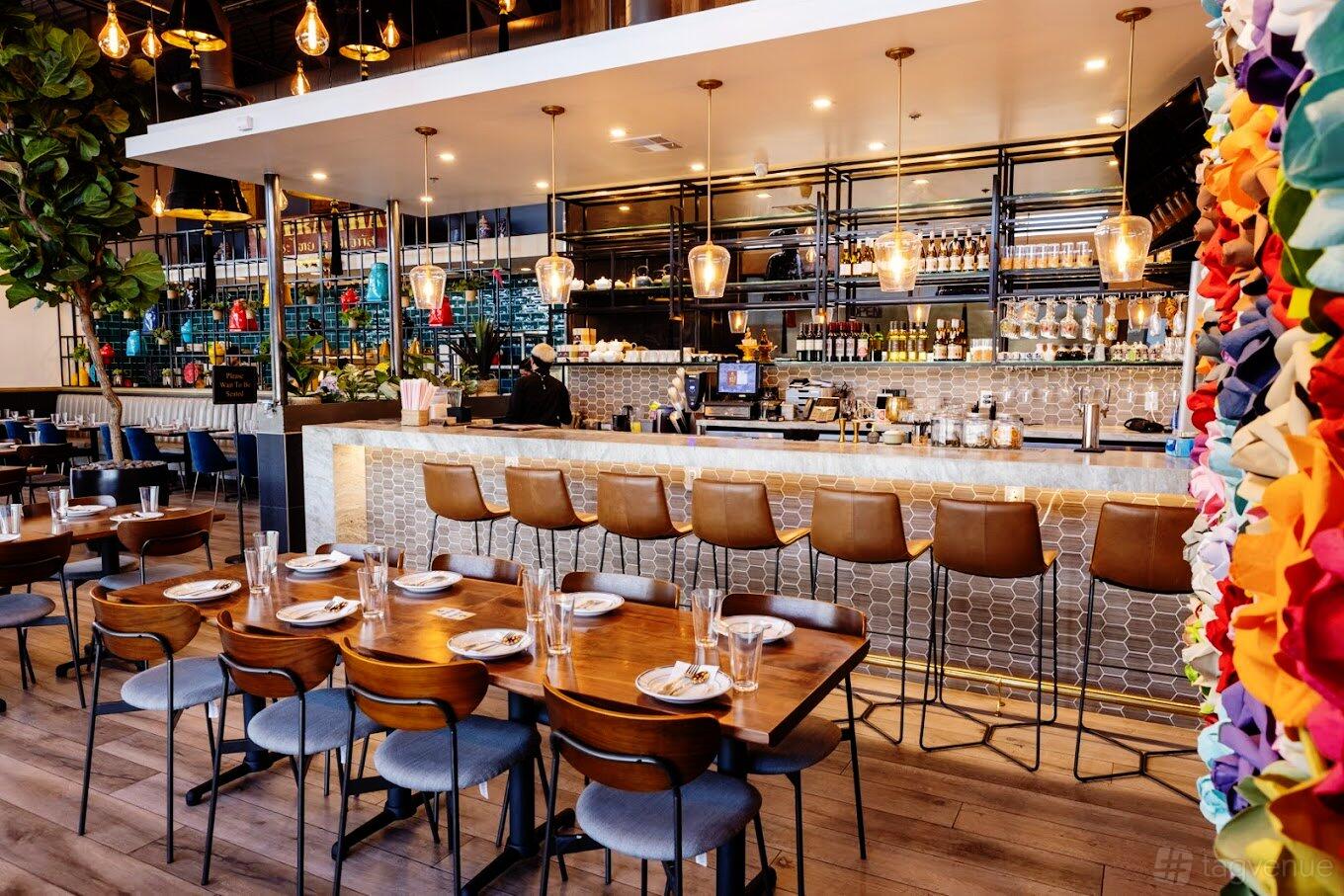 A restaurant with a long backlit bar, hanging pendant lights, and wooden dining tables at Weera Thai - Rainbow.