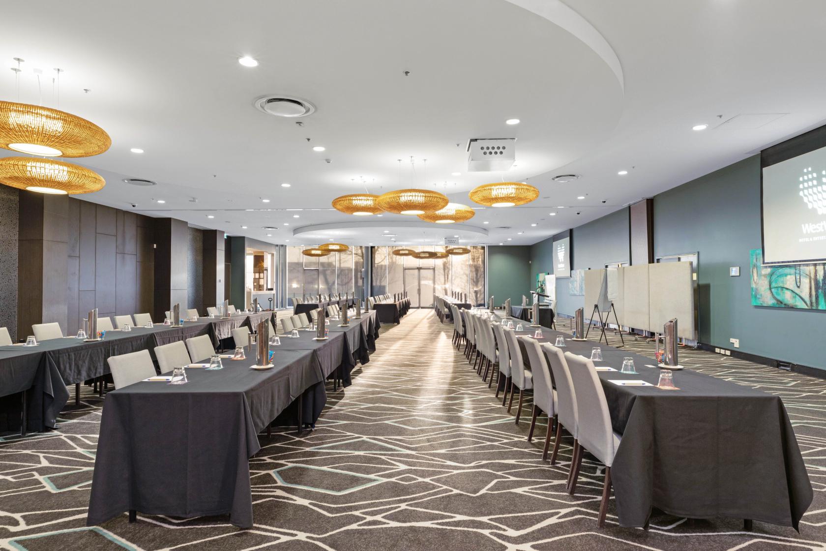 A ballroom with geometric carpet, long tables set classroom-style, and round pendant lights at WestWaters Hotel & Entertainment Complex.