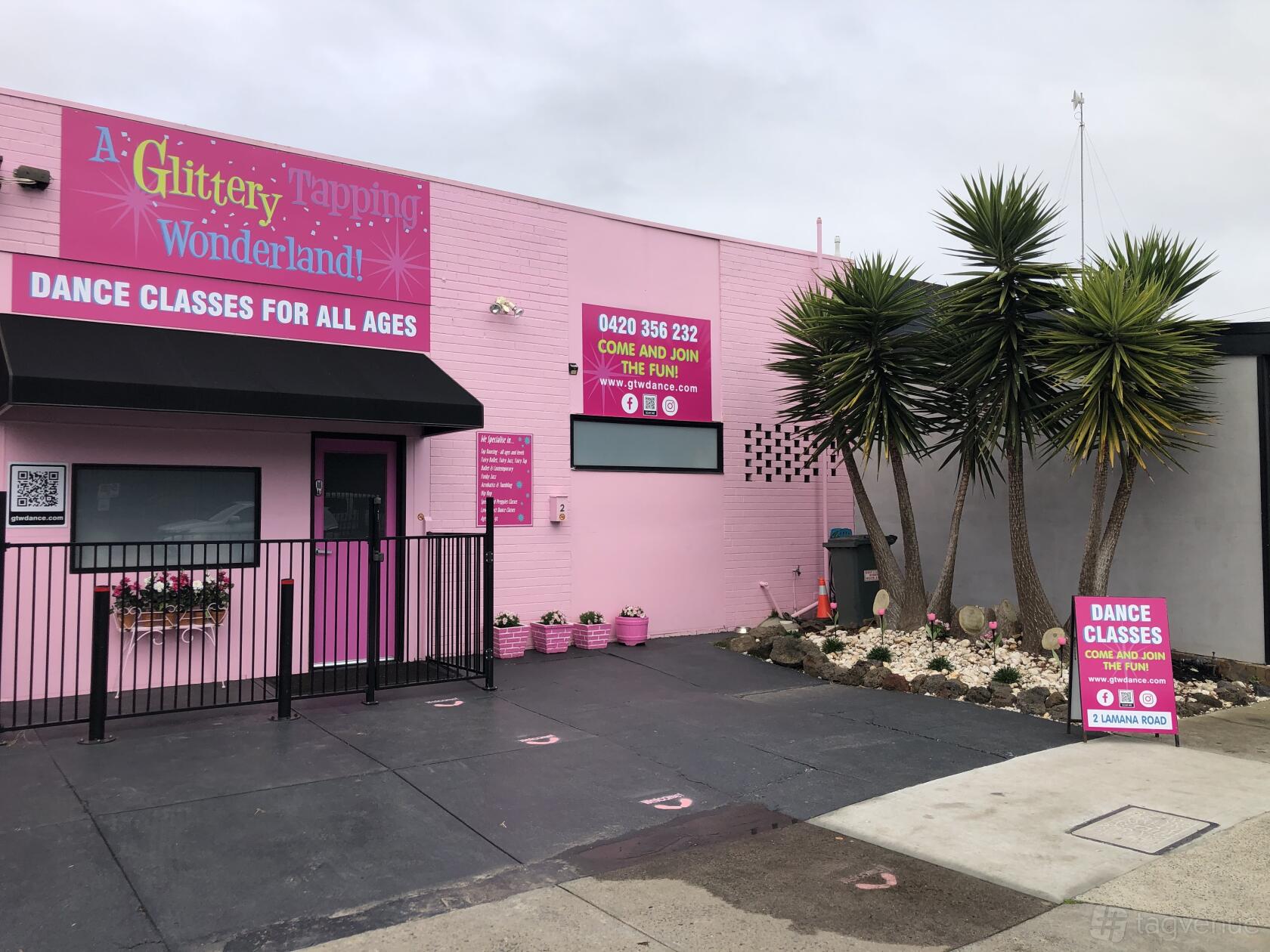 A pink dance studio exterior with bold signage, large windows, and yucca plants at Glittery Tapping Wonderland.