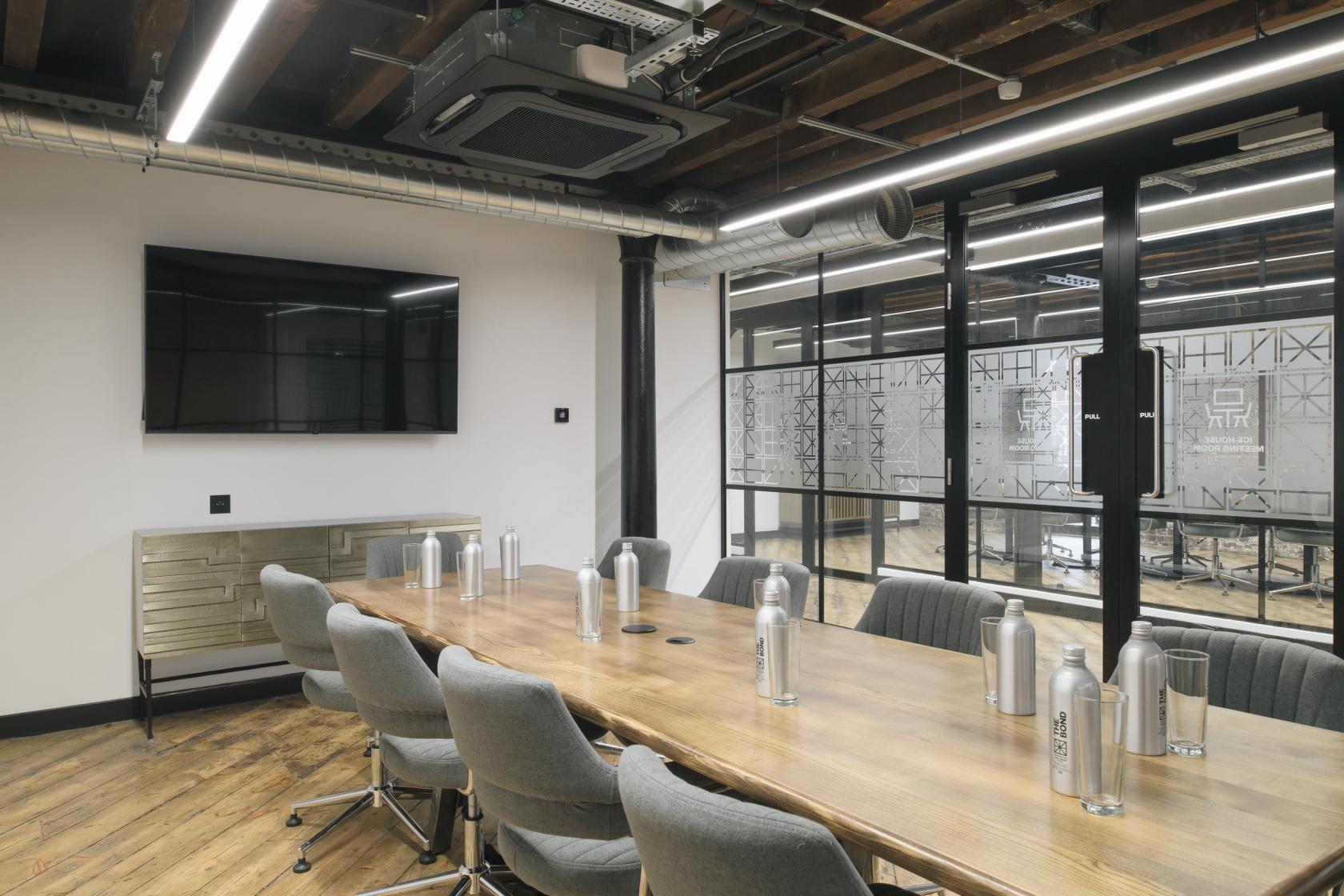 A boardroom with a long wooden table, gray upholstered chairs, exposed beams, and a large wall-mounted screen at The Bond.