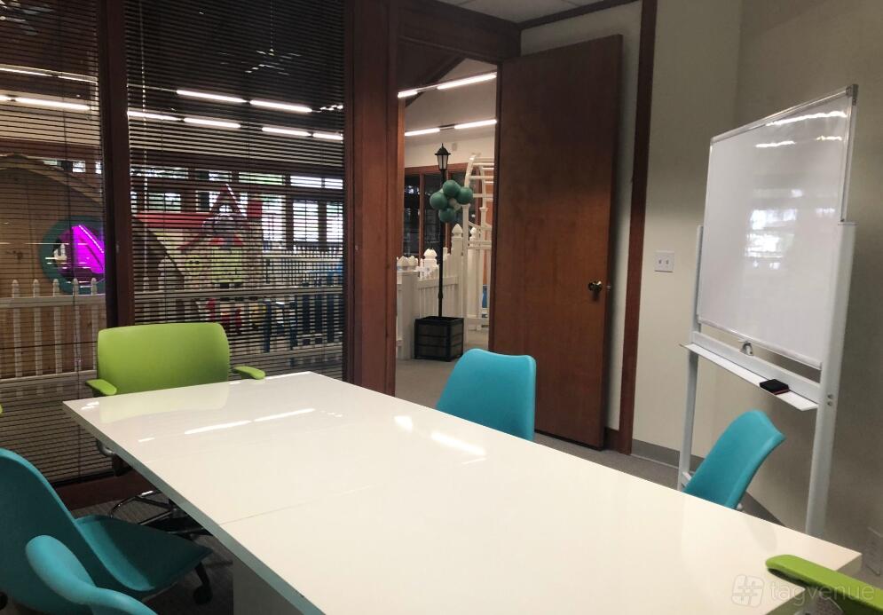 A meeting room with a white table, colorful chairs, glass wall panels, and a mobile whiteboard at Fandory Factory.