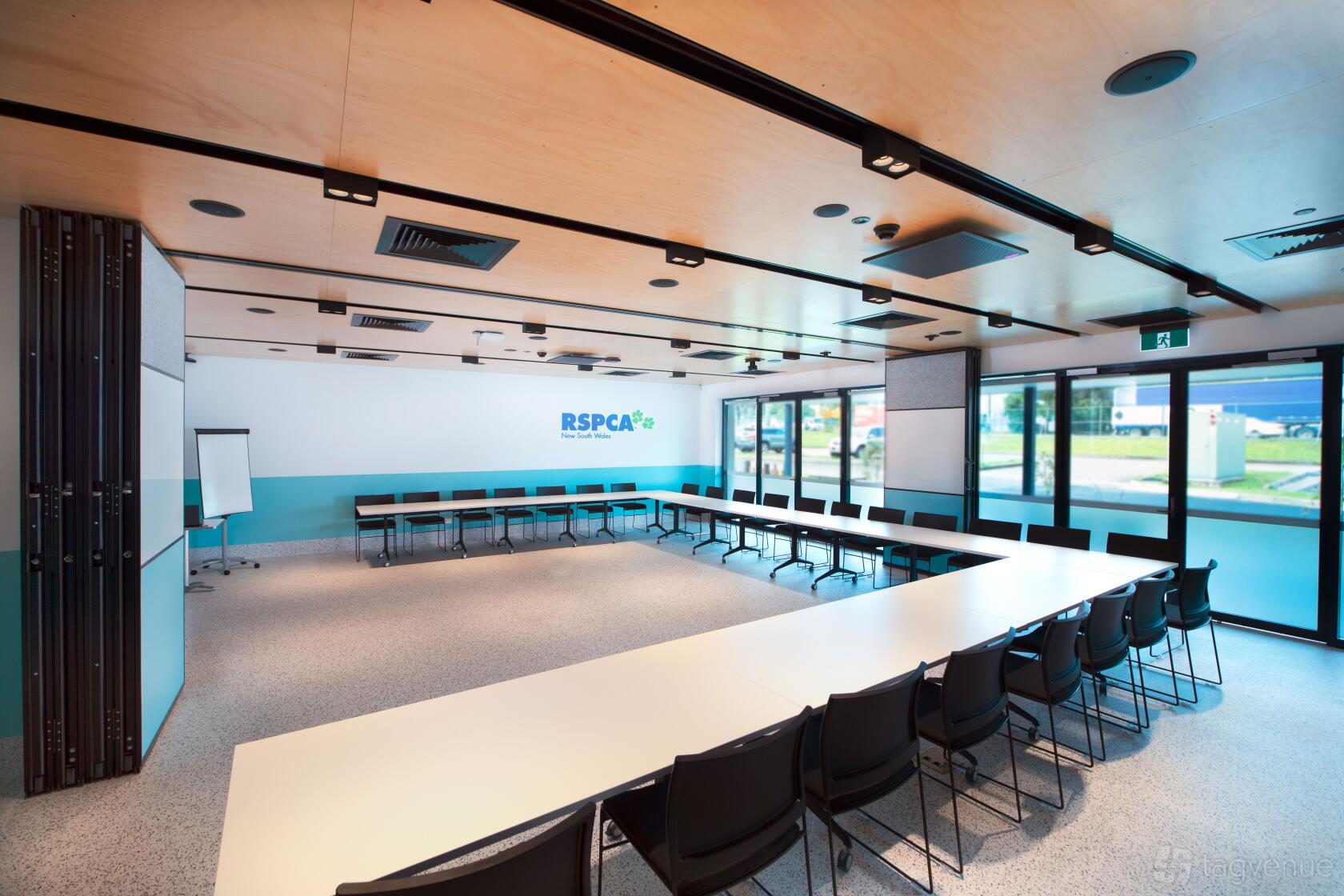 A meeting room with U-shaped white tables, black chairs, natural light, and a flip chart at RSPCA NSW Sydney Education Centre.