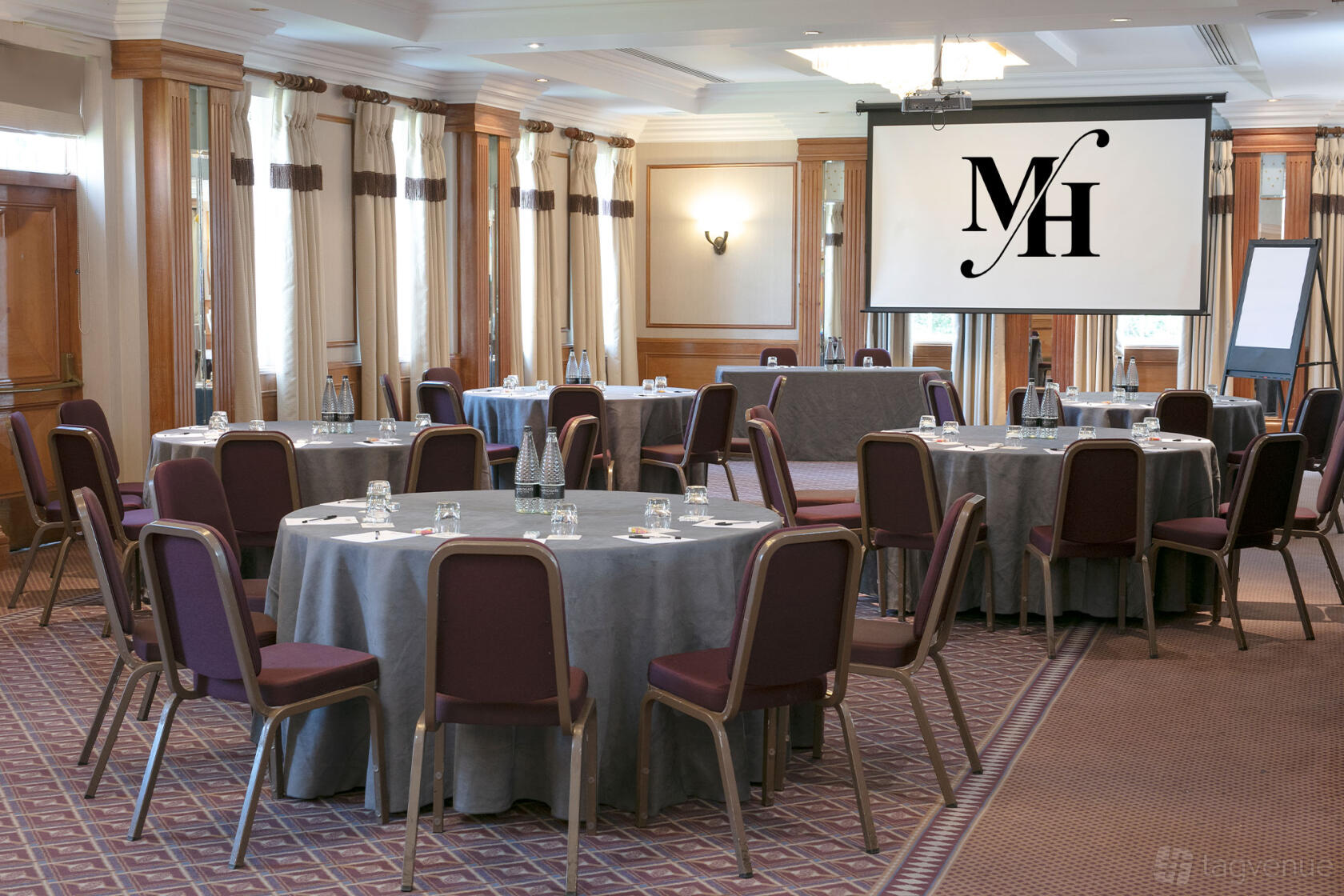 A hotel meeting room with round tables covered in gray linens, mauve chairs, and a ceiling-mounted projector at Best Western Plus Manor Hotel.