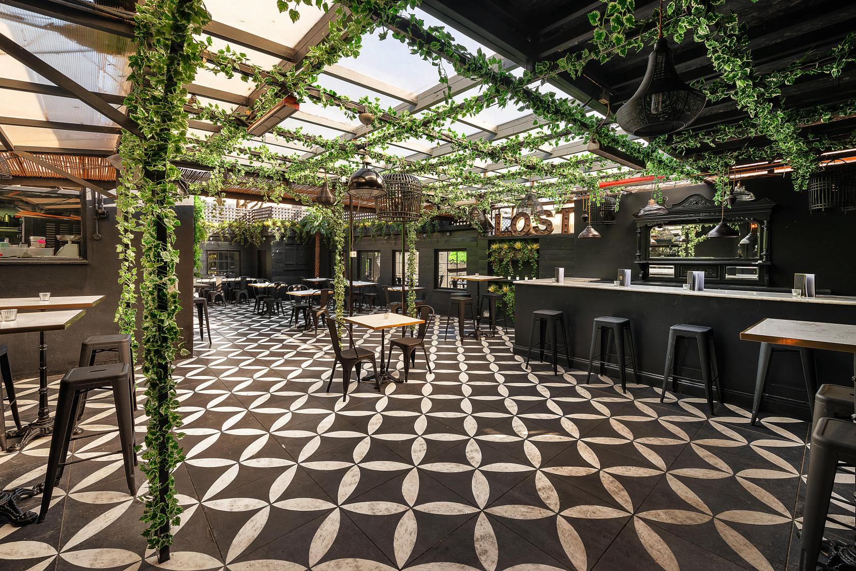 An outdoor cocktail bar with geometric tiled flooring, hanging greenery, and a covered roof at Lost Society (Battersea SW11).