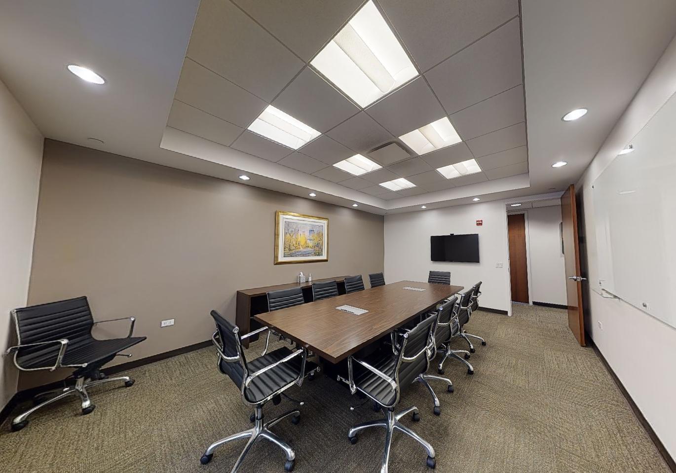 A conference room with a long wooden table, black ergonomic chairs, a wall-mounted TV, and framed artwork at Corporate Suites - Lexington Avenue.