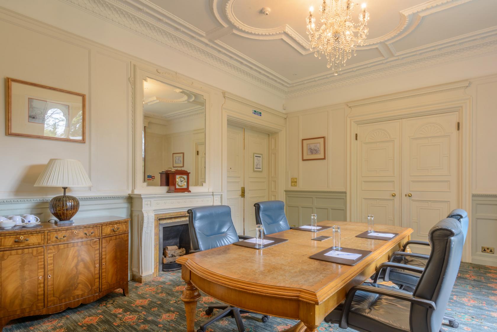 An event space with a wooden meeting table, black leather chairs, chandelier, and ornate ceiling at Warren House Hotel.